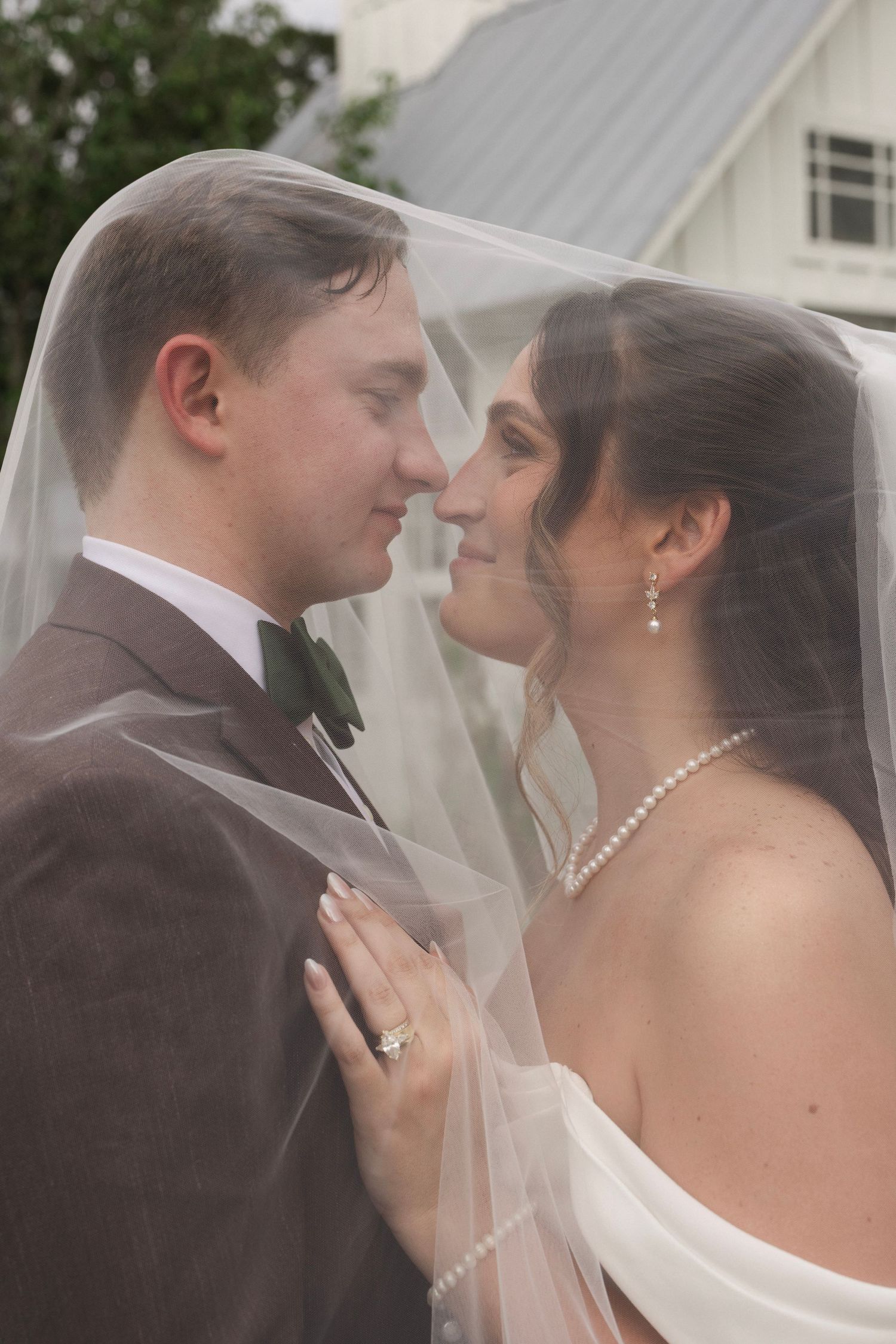 Bride and groom share an intimate moment under a wedding veil, bride wearing pearl jewelry and engagement ring.