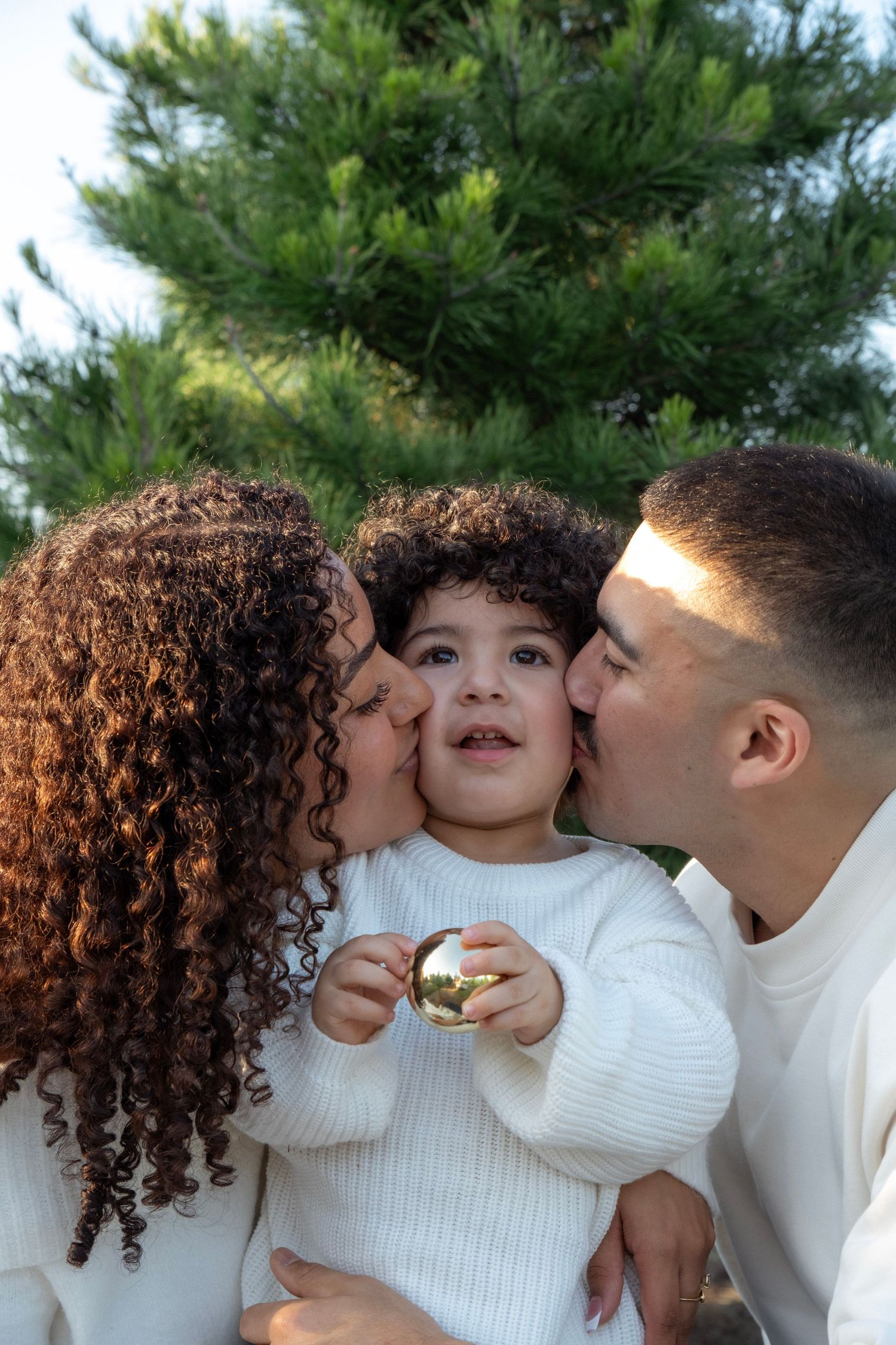 Parents kissing their curly-haired child on the cheeks outdoors near green pine trees.