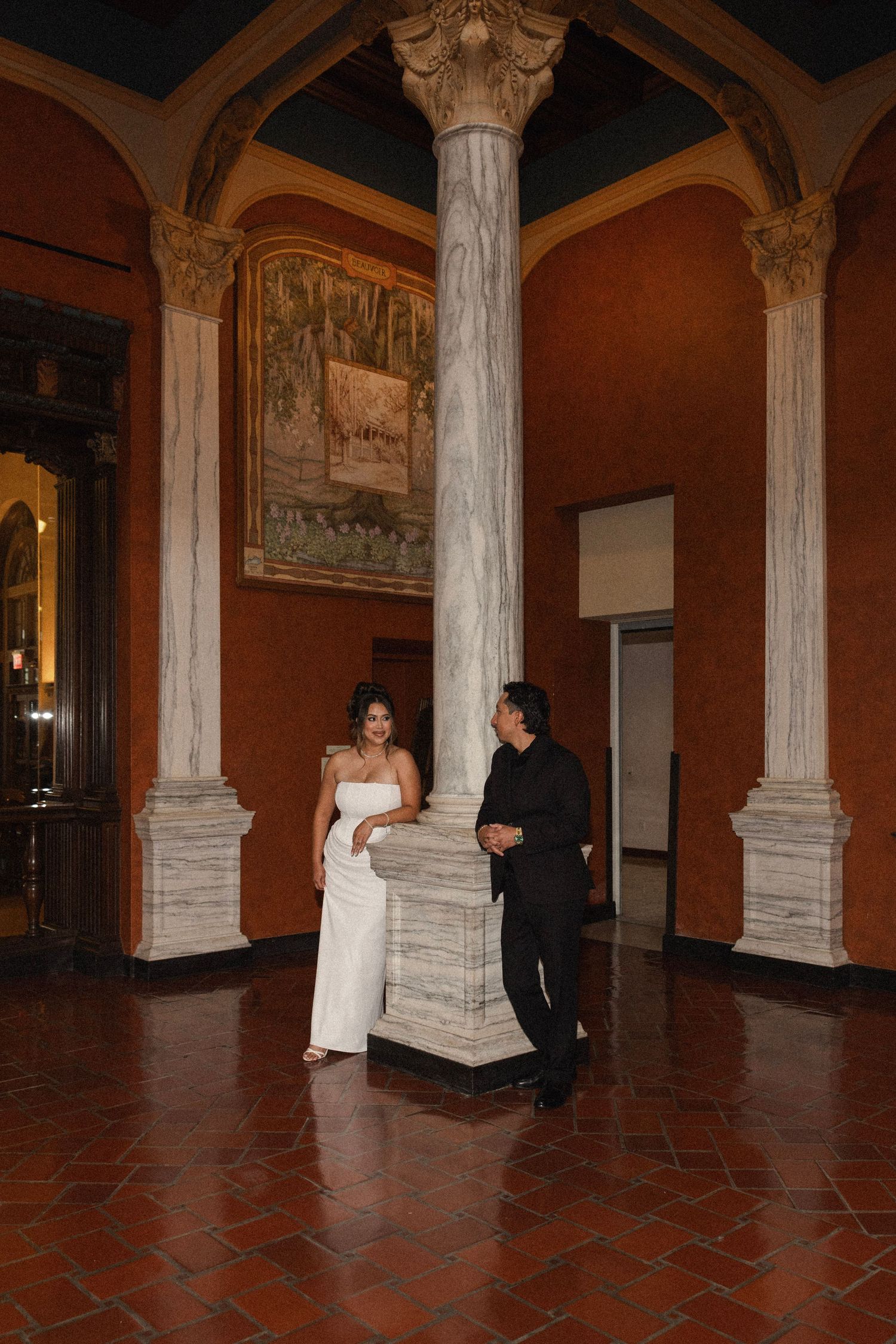 Couple in elegant attire standing by marble columns in an ornate hall with terracotta floors and murals.