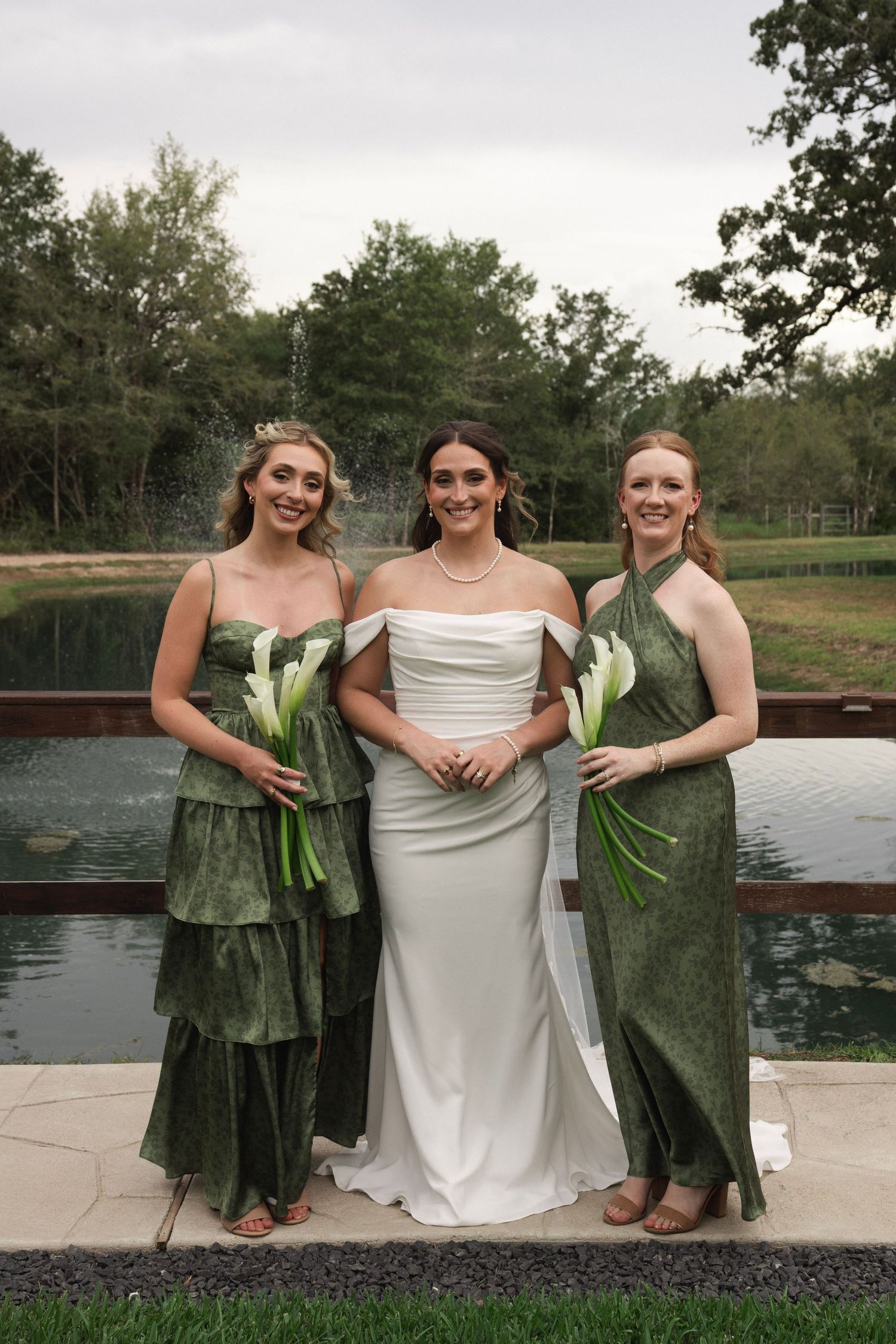 Bride in white off-shoulder gown with two bridesmaids in green dresses holding calla lilies by a pond.
