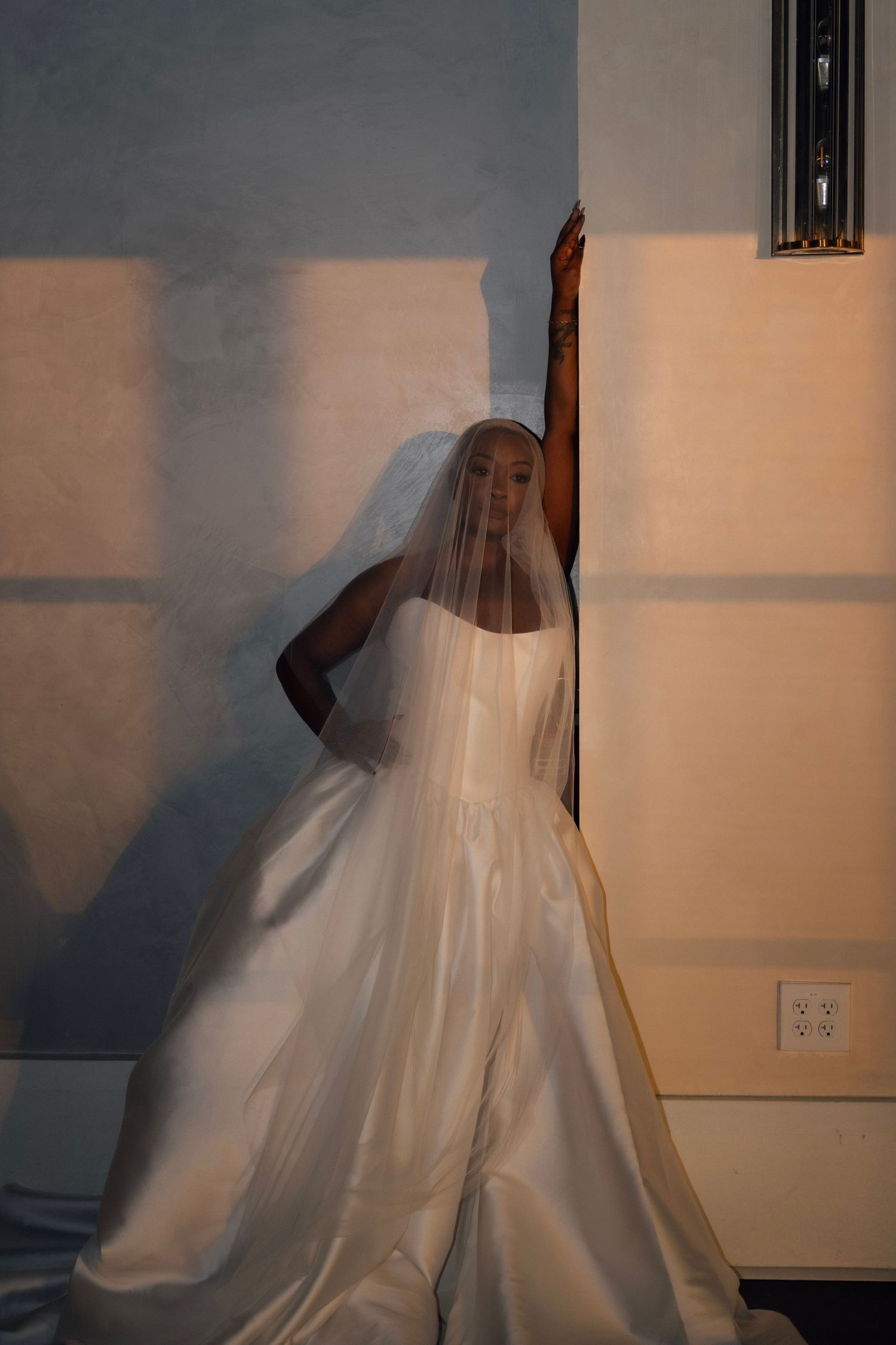 Bride in white ball gown and long veil leans against a wall in dramatic golden and blue light.