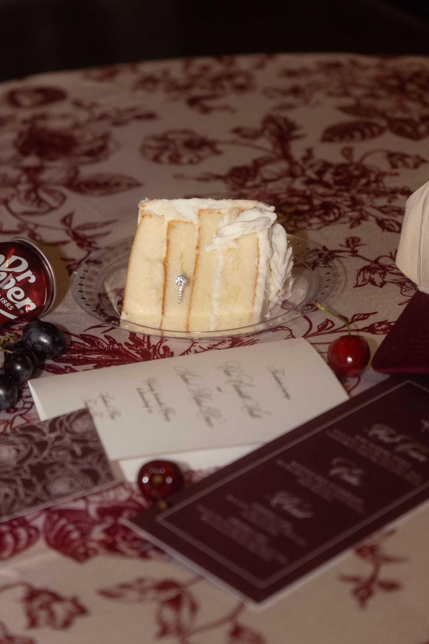 Wedding cake slice displayed on a table with menus, red ornaments, and a burgundy floral toile tablecloth.