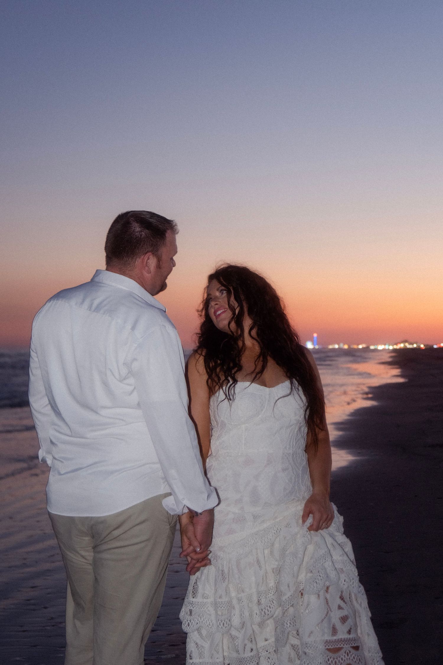 Couple holding hands on beach at sunset, woman in white lace dress and man in white shirt smiling at each other.