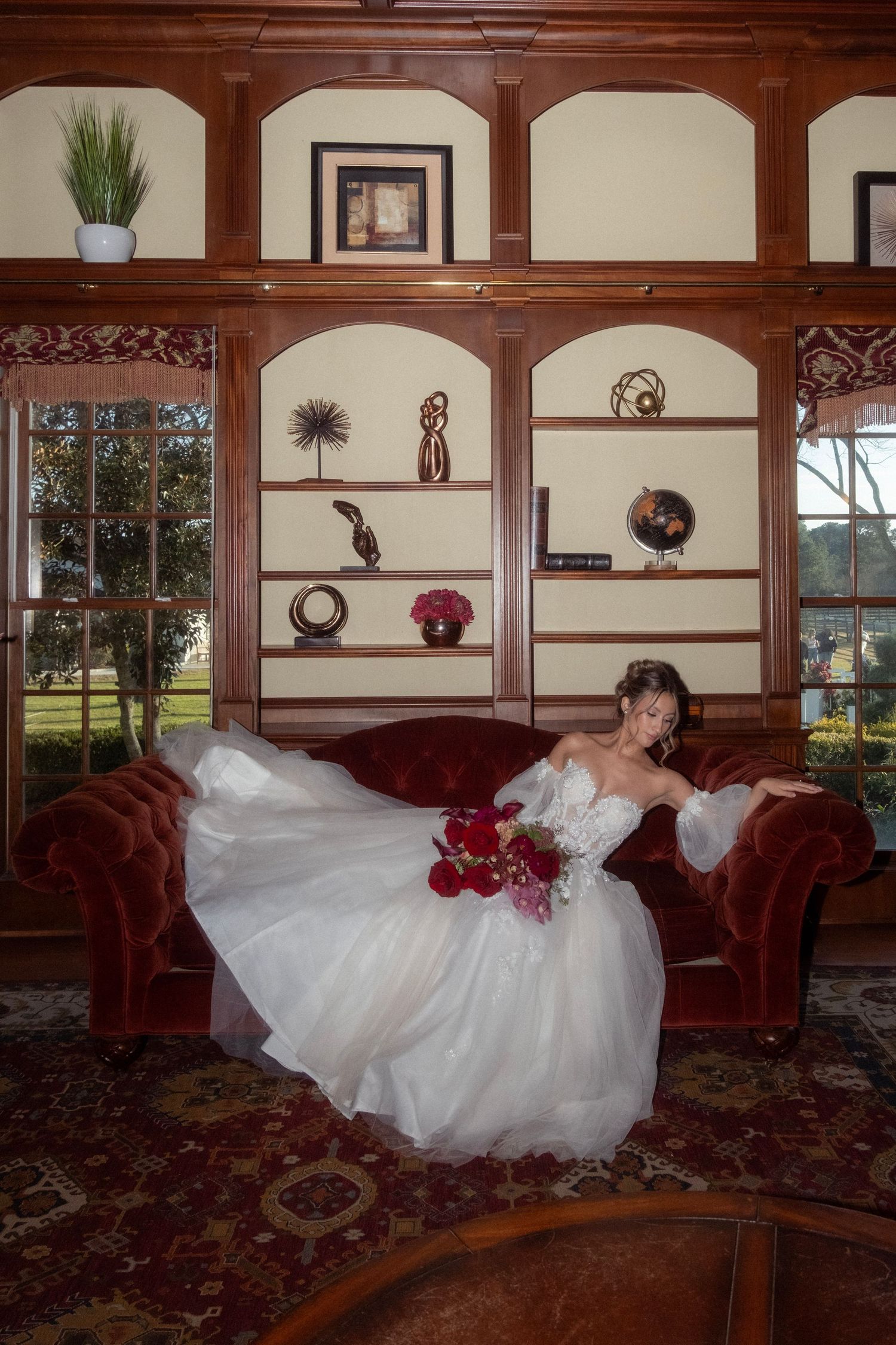 Bride in a flowing white gown sits on a red velvet sofa holding a bouquet.