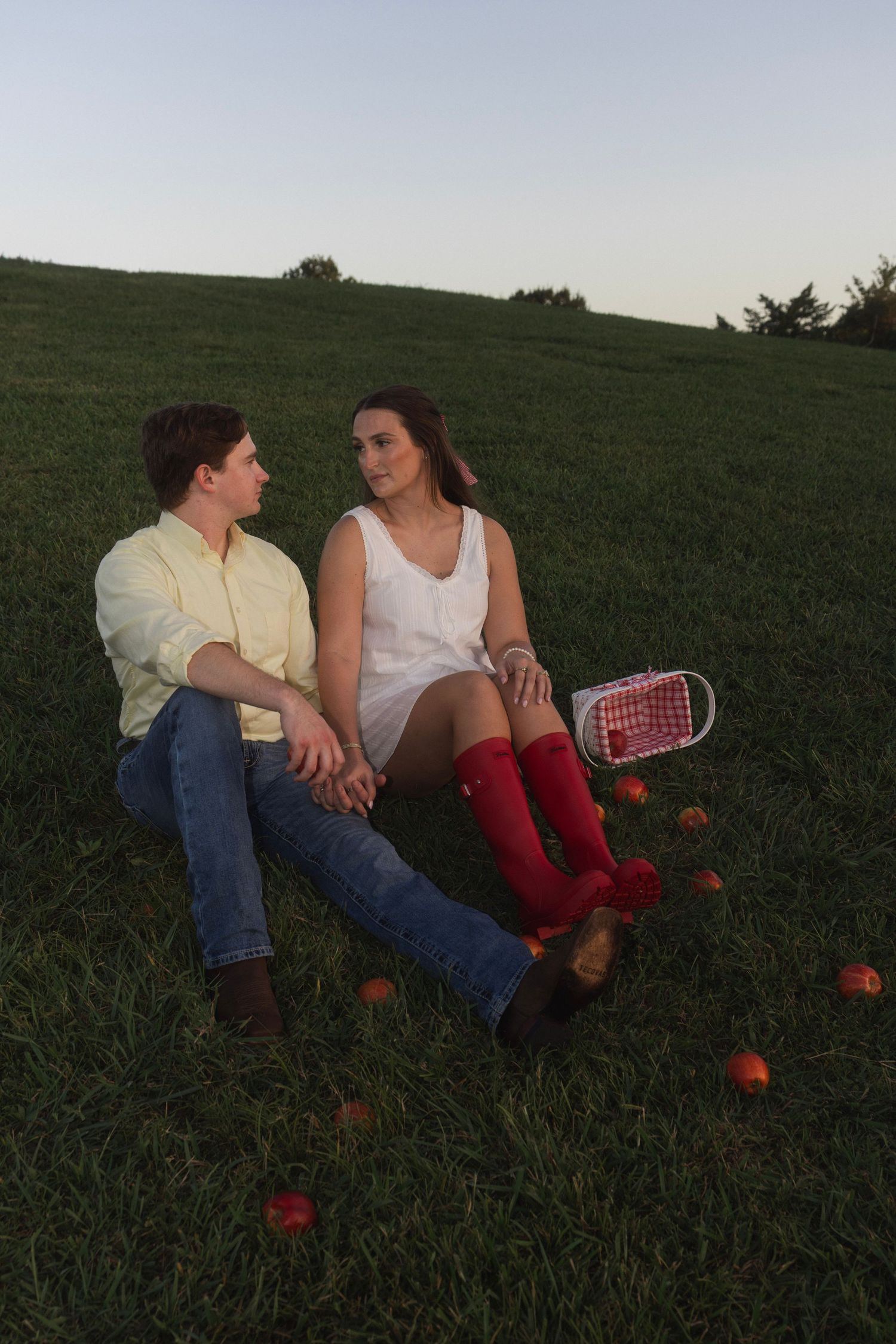 A young couple sits on a grassy hill with scattered apples and a basket, gazing at each other at dusk.
