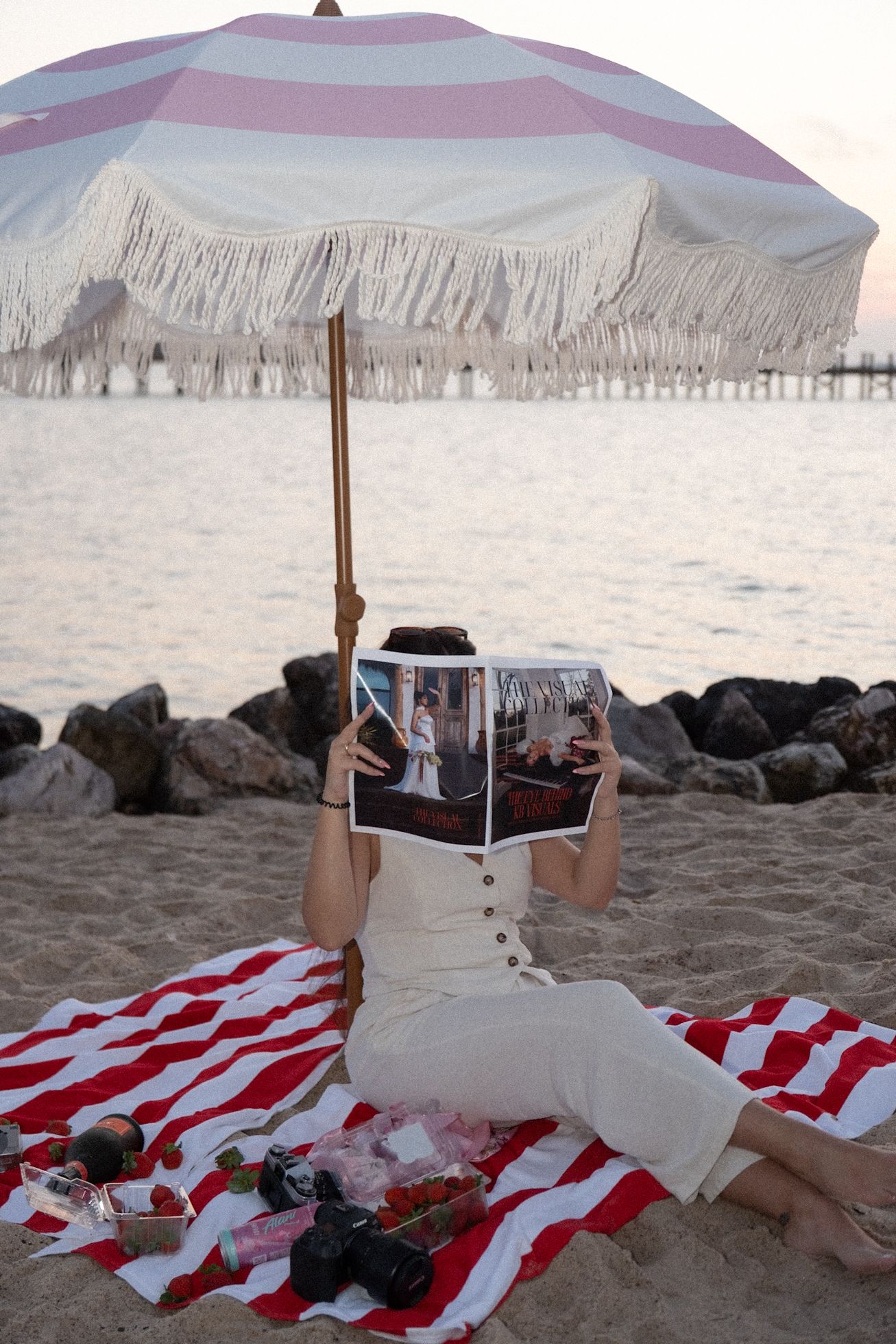 Woman in white dress sits under striped fringed umbrella on beach holding wedding photo album.