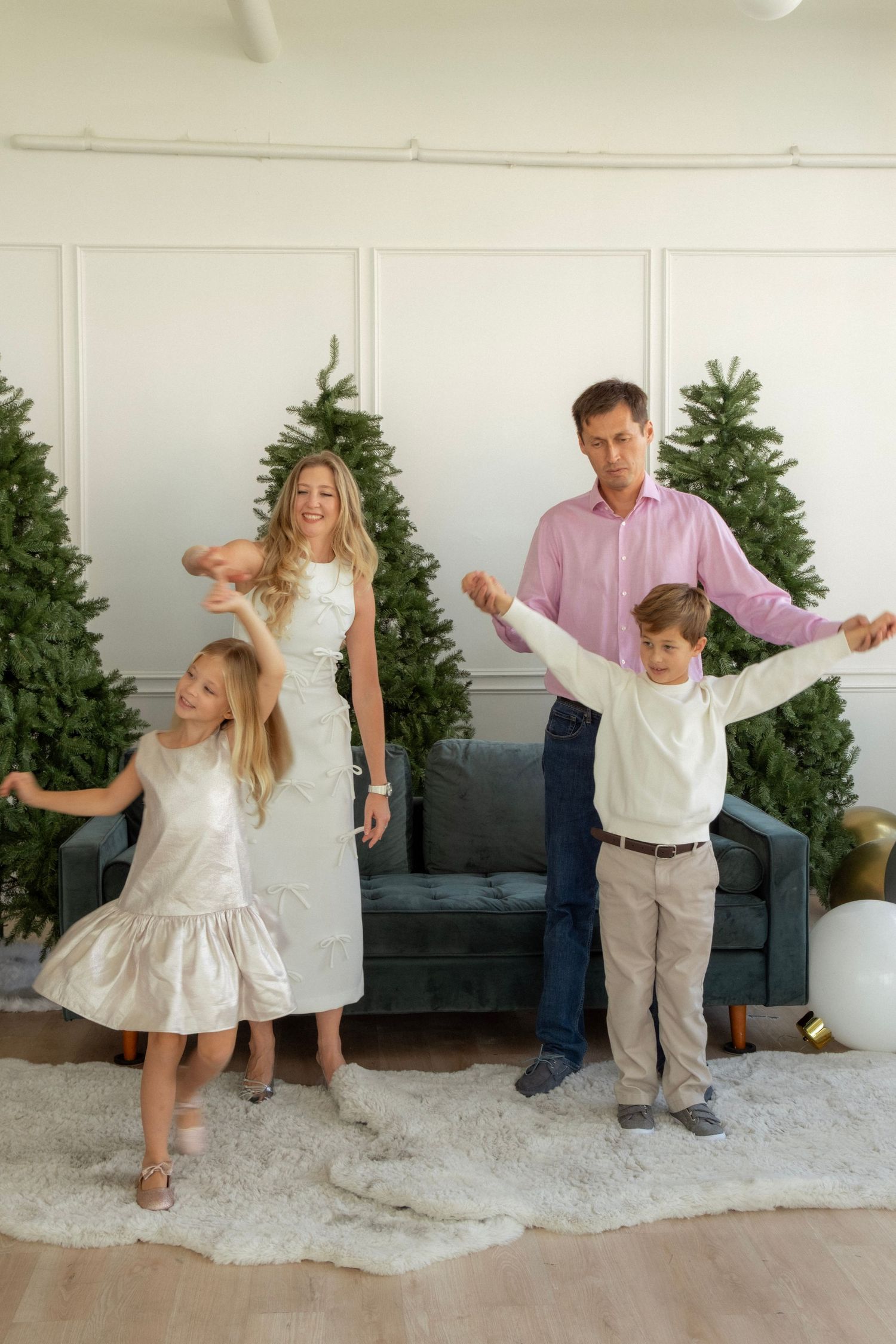 Family of four dancing joyfully in a white living room decorated with Christmas trees, wearing elegant holiday attire.