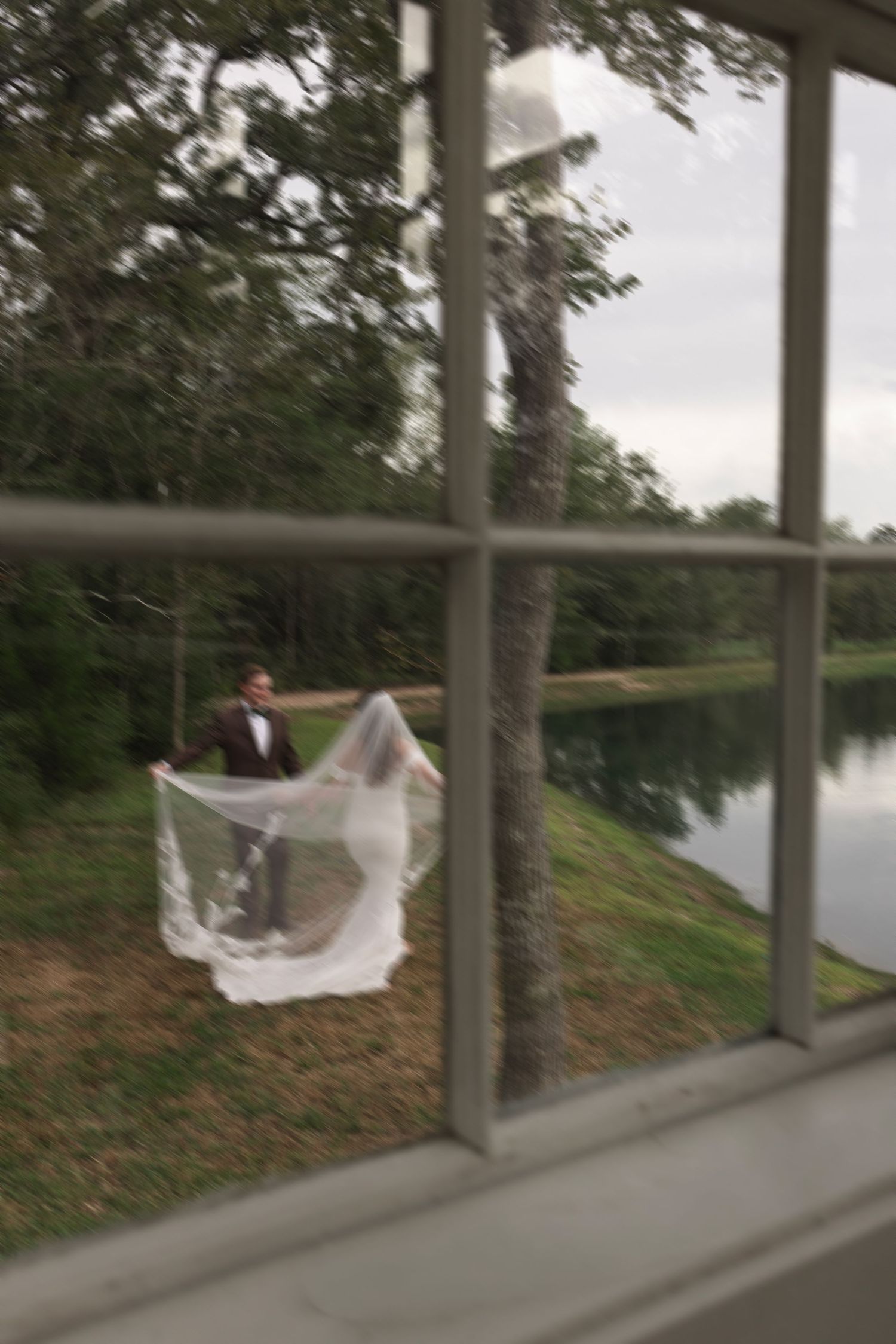 Bride and groom sharing a romantic moment outdoors near a serene lake, viewed through a window, with veil flowing in the breeze.