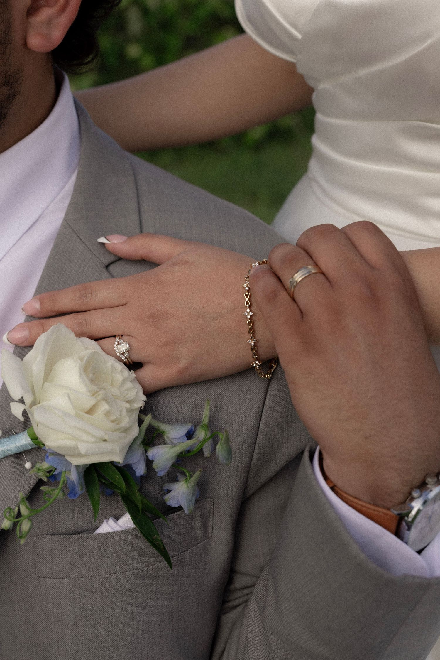 Close-up of newlyweds hands showing wedding rings with white rose boutonniere on grooms gray suit lapel.