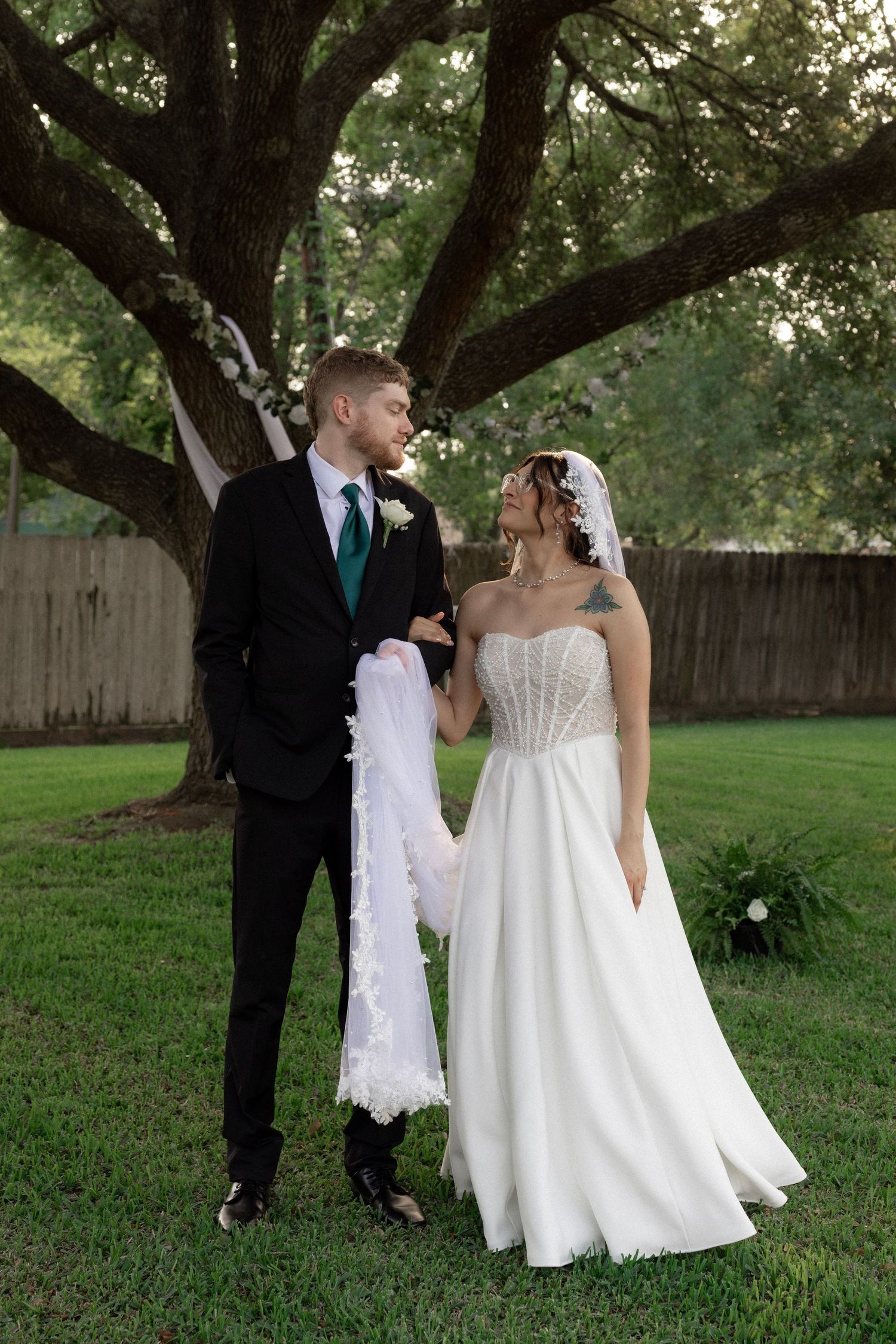 Bride and groom stand together under a large oak tree in a lush green backyard.