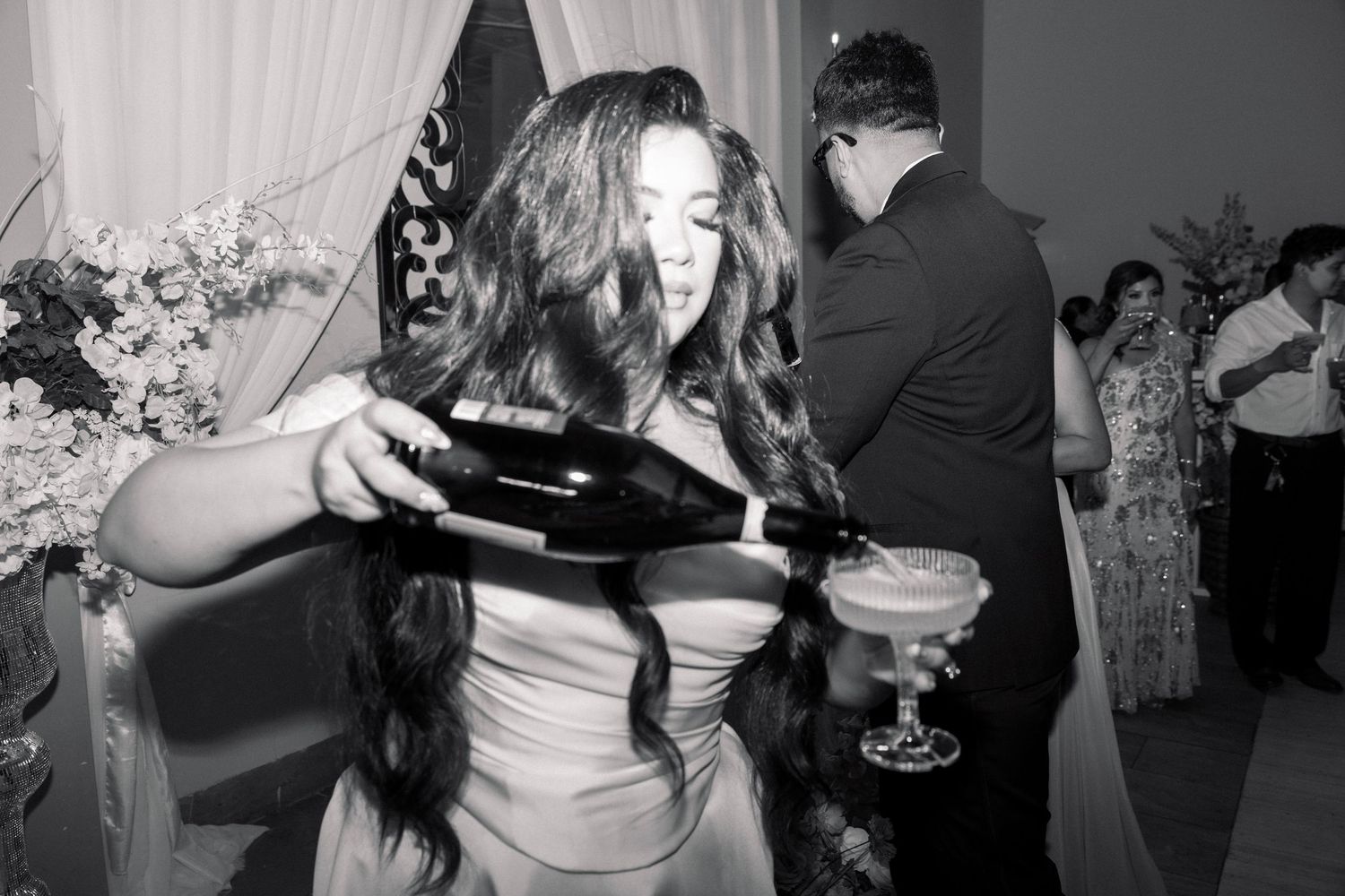Black and white photo of bride laughing while pouring champagne bottle at wedding reception.