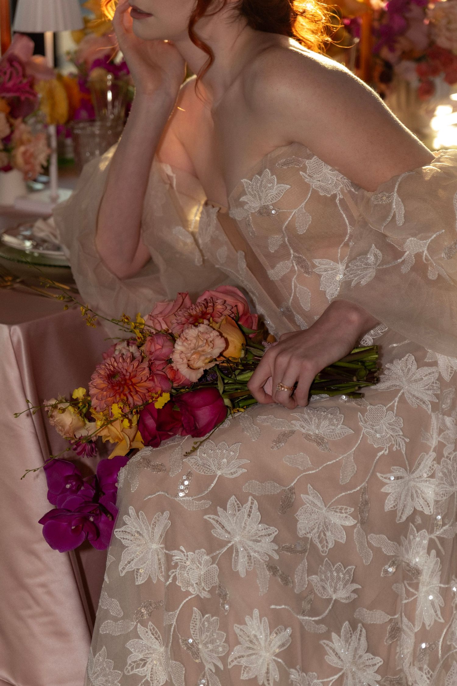 Bride in floral lace gown holds vibrant pink and orange bouquet while seated at decorated reception table.