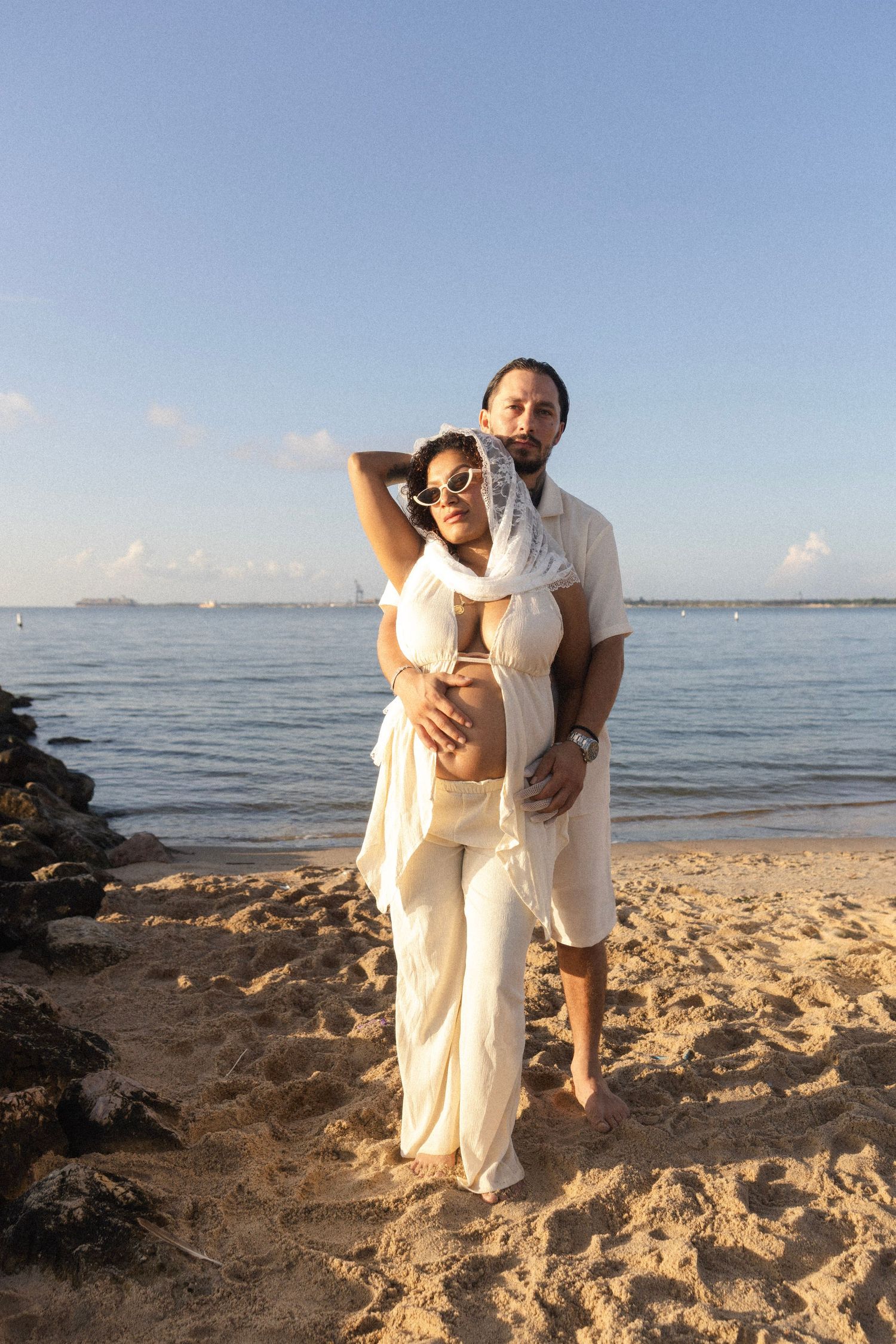 Couple in white outfits embracing on a sandy beach with calm ocean waters and clear blue sky behind them.