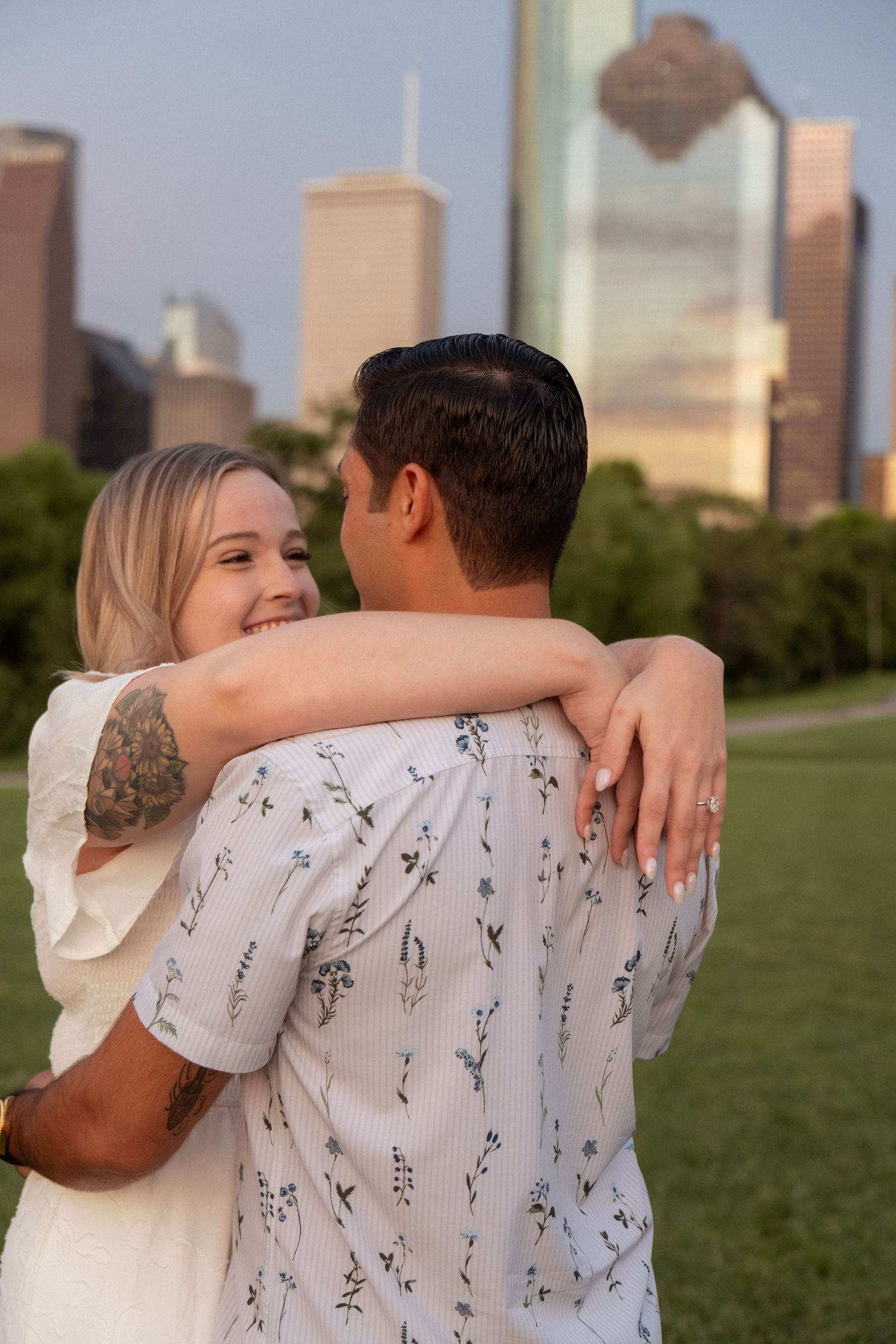 A couple embraces in a park with the Houston skyline at sunset, the woman smiling with a floral tattoo on her arm.