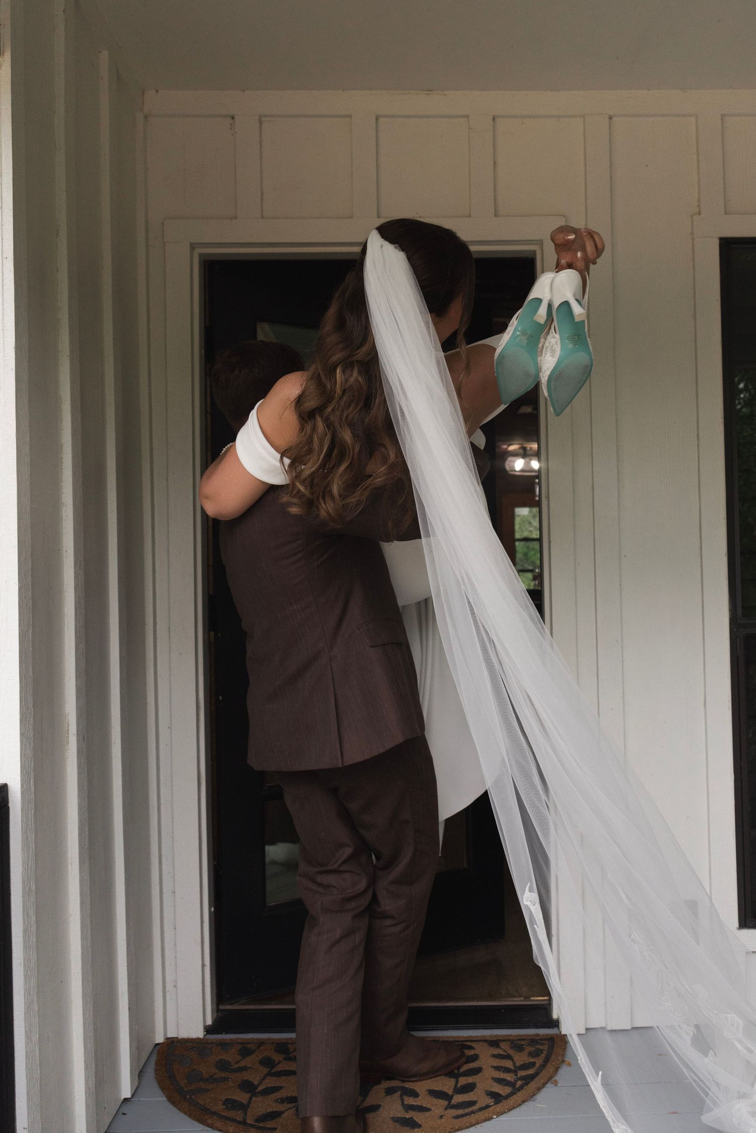 Bride in wedding veil being helped by groomsman in brown suit at doorway, holding teal shoes.