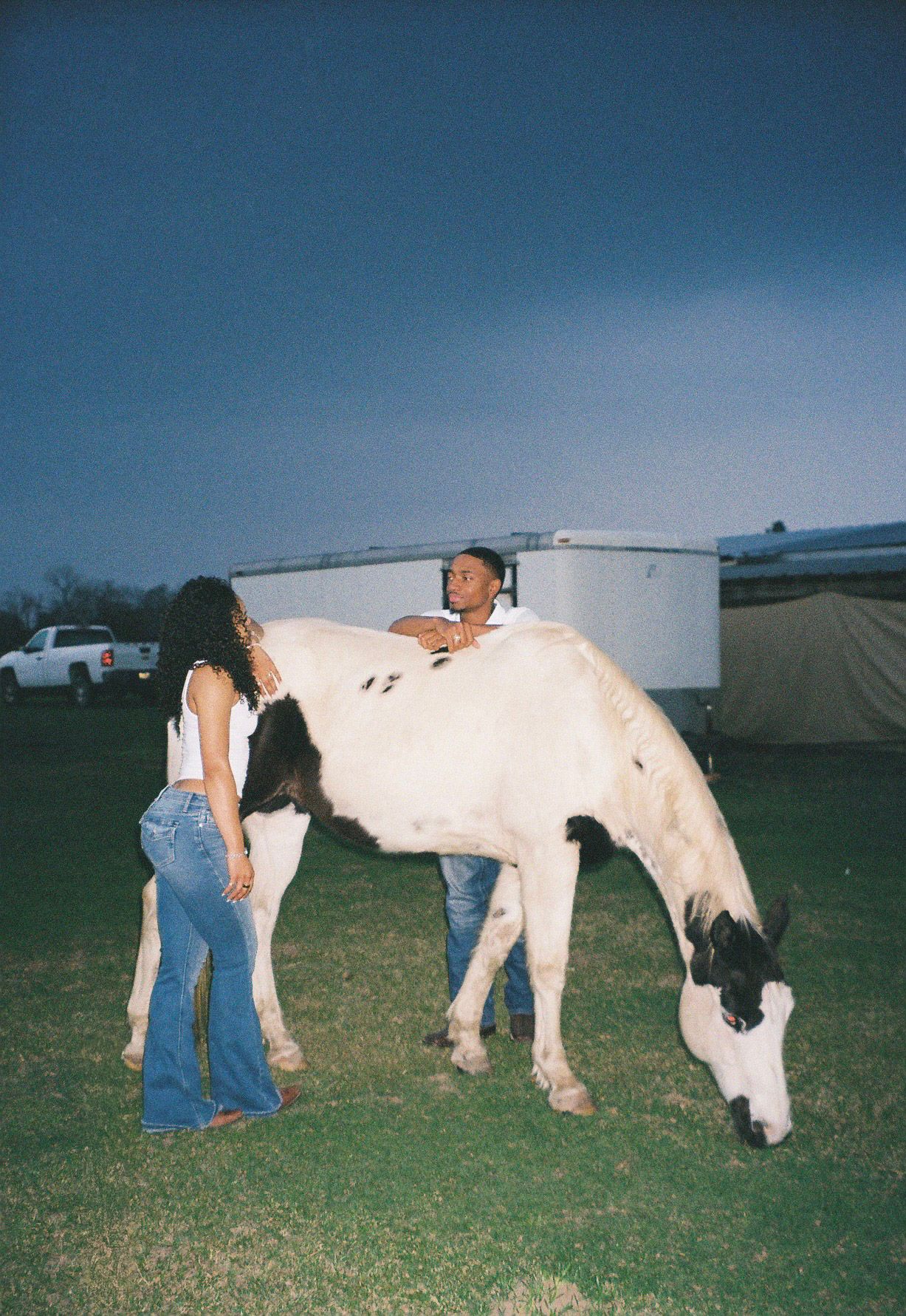 Couple posing with a black and white paint horse on a farm at dusk.