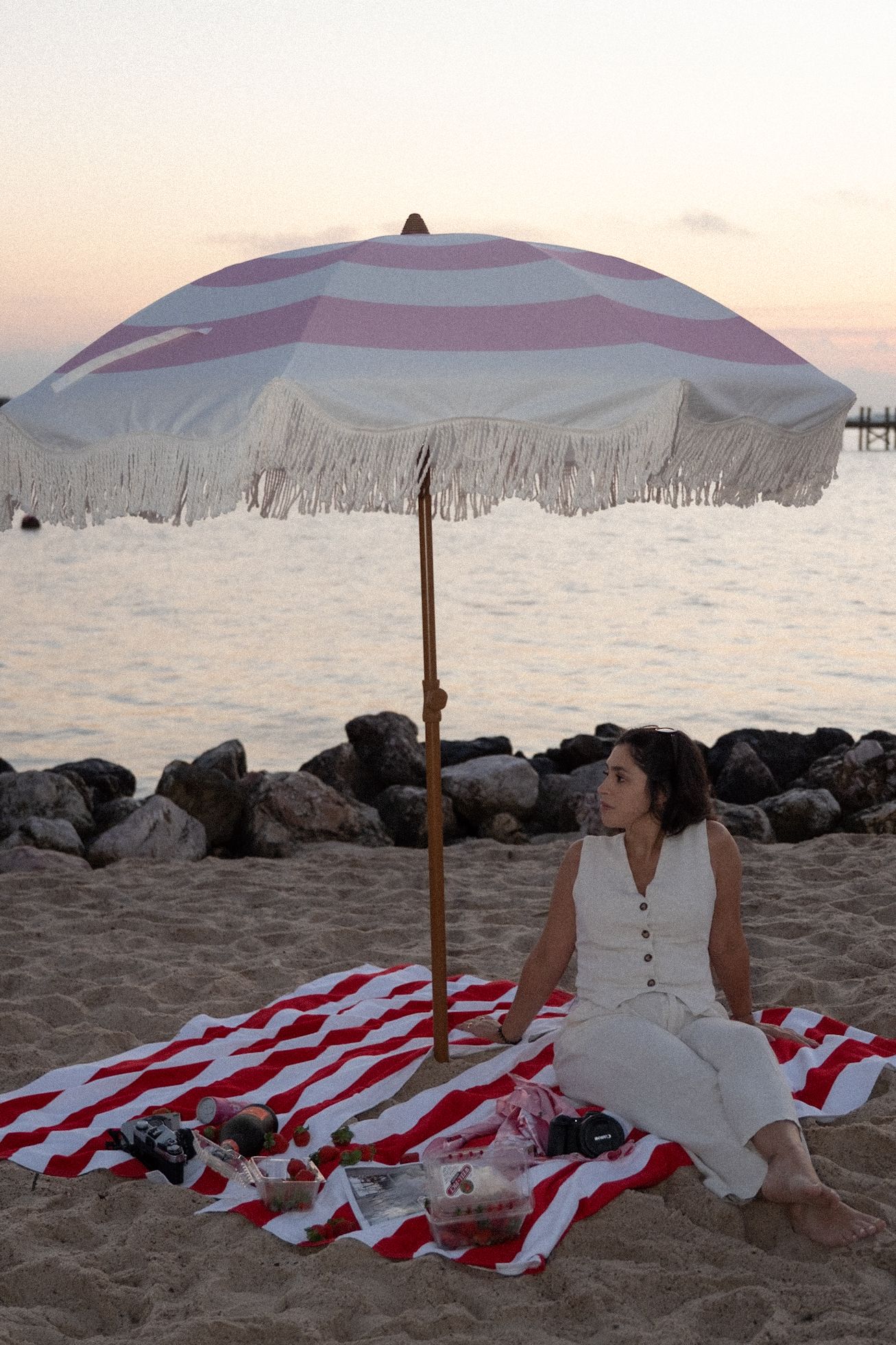 Woman in white dress sits on striped blanket under fringed beach umbrella at sunset by the ocean.