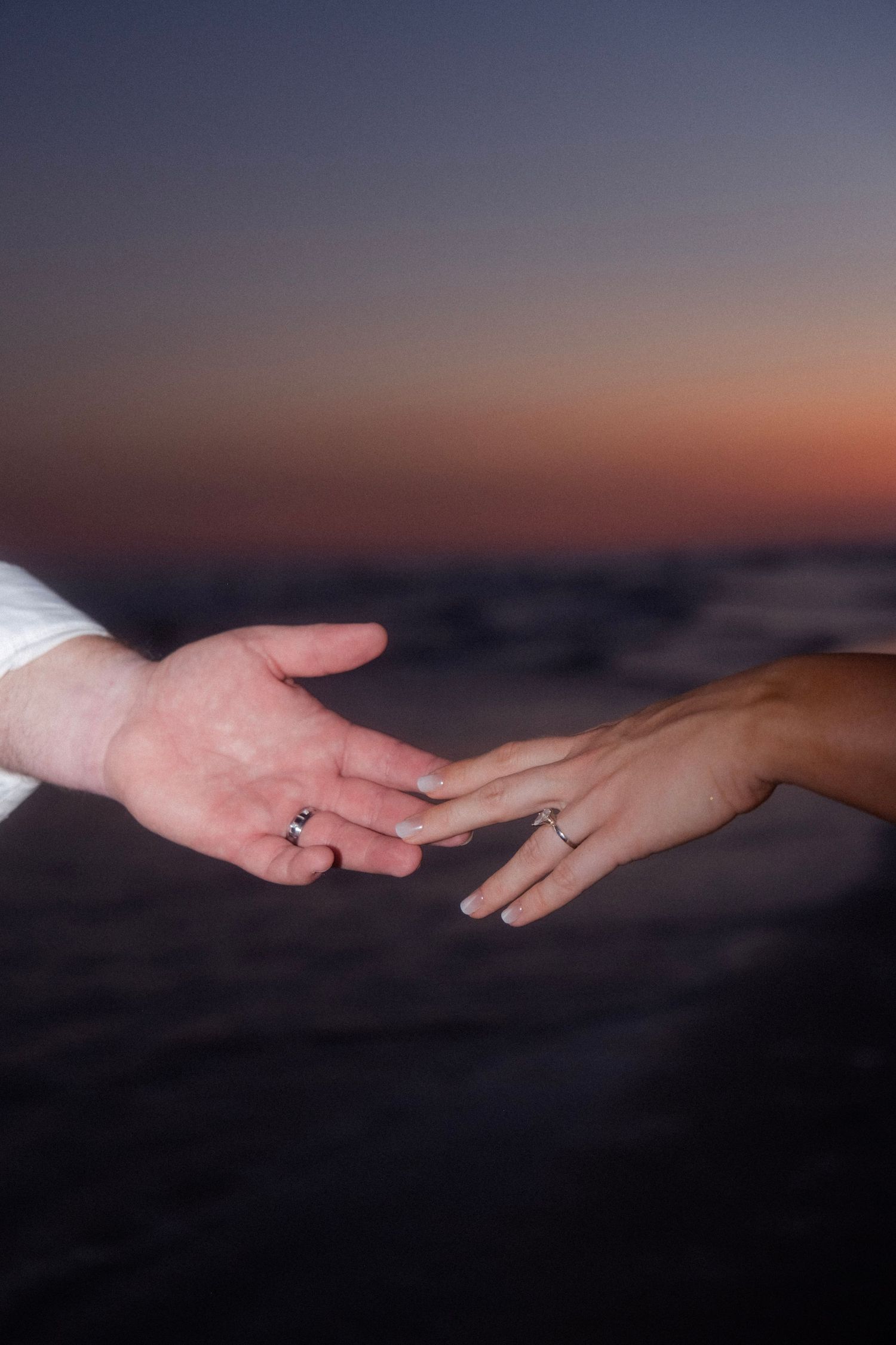 Newlywed couple reaching hands toward each other with wedding rings at sunset beach.
