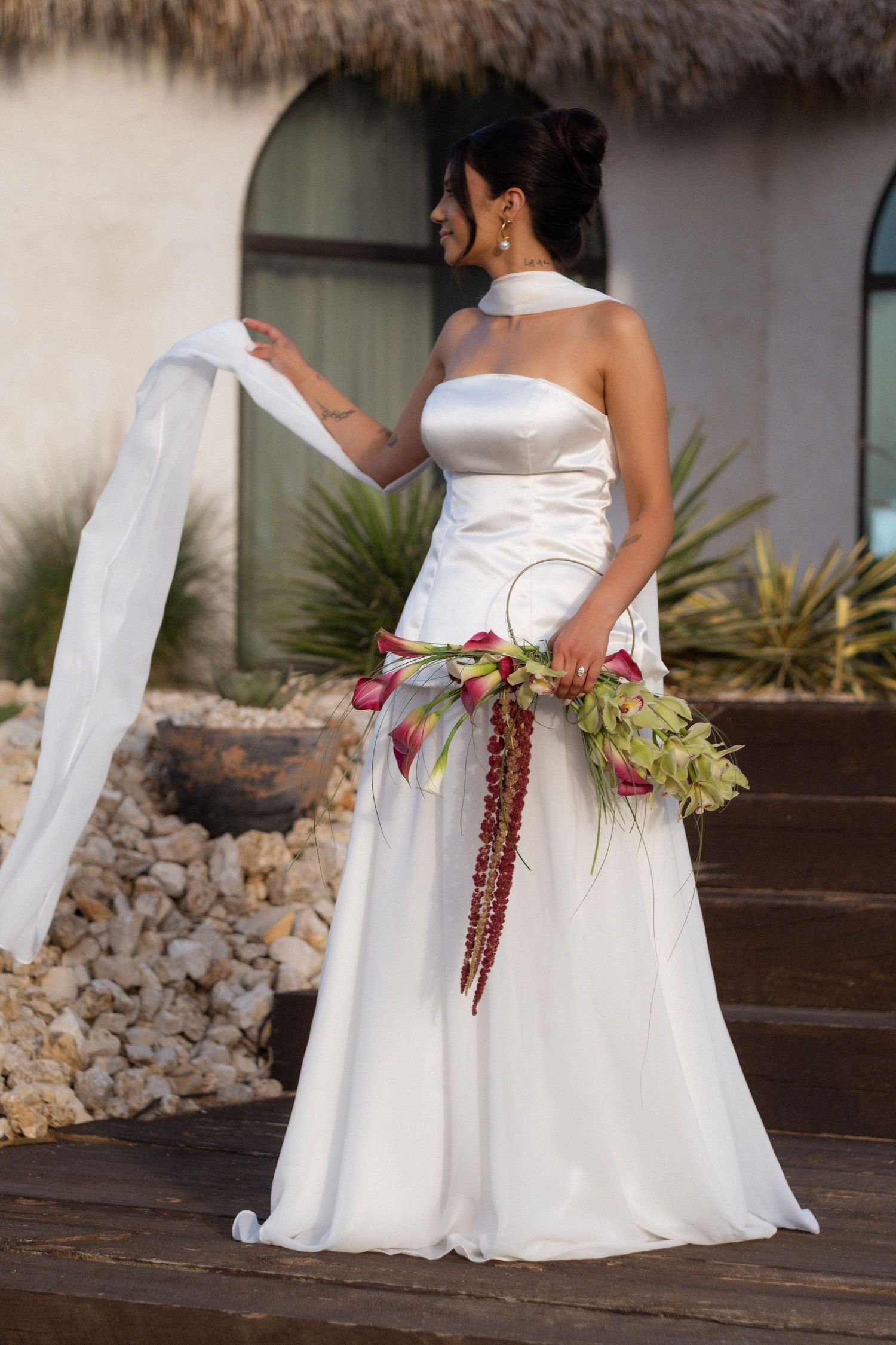 Radiant bride in strapless satin gown holding tropical bouquet with flowing veil at a resort in Mexico.