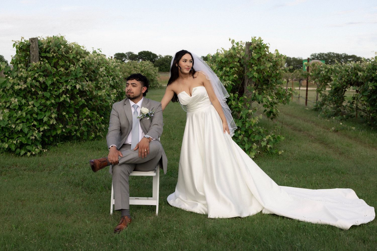 Bride in white gown and groom in gray suit pose in lush vineyard for elegant outdoor wedding portrait.