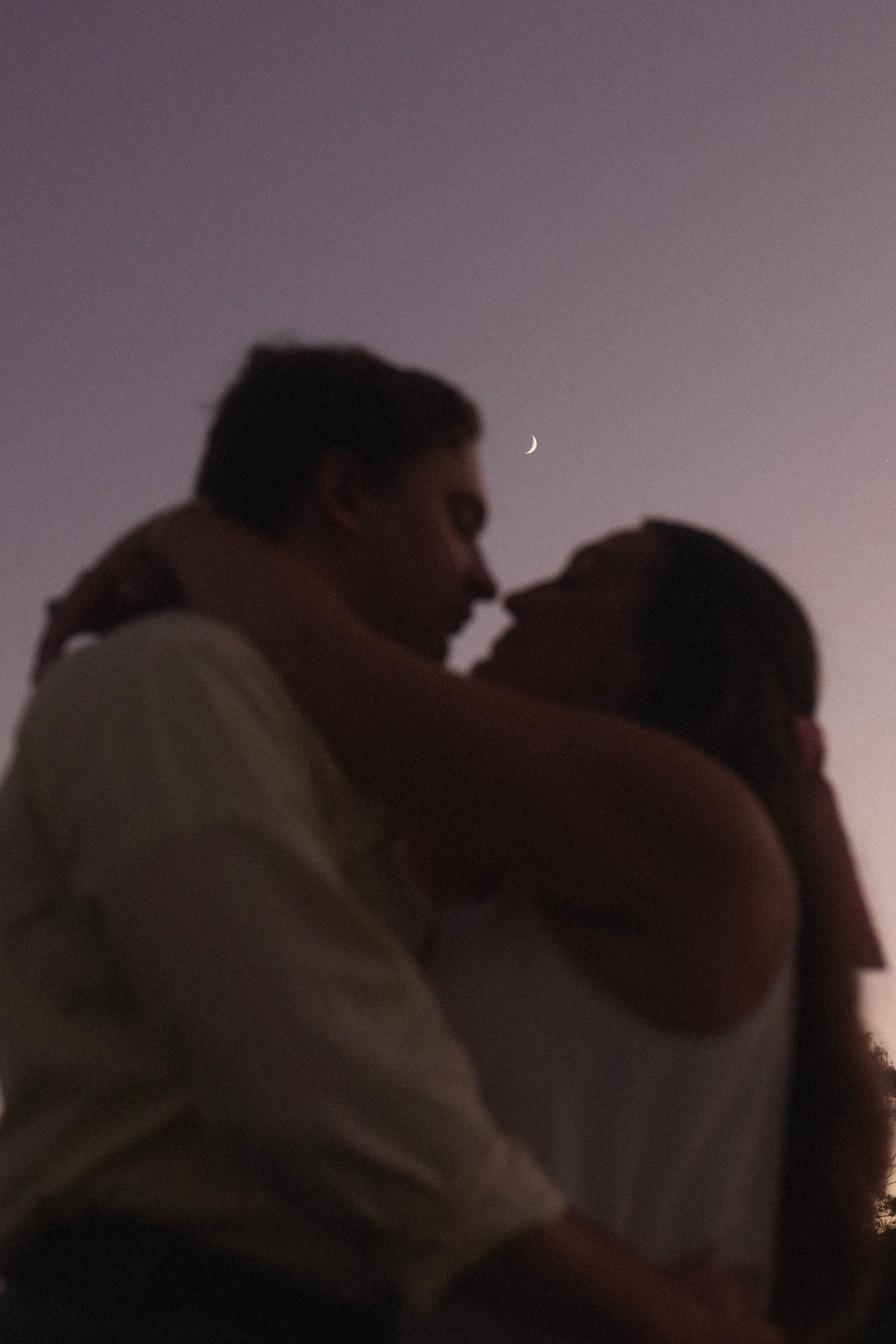 Romantic couple sharing a kiss at dusk with a crescent moon glowing in the purple twilight sky.