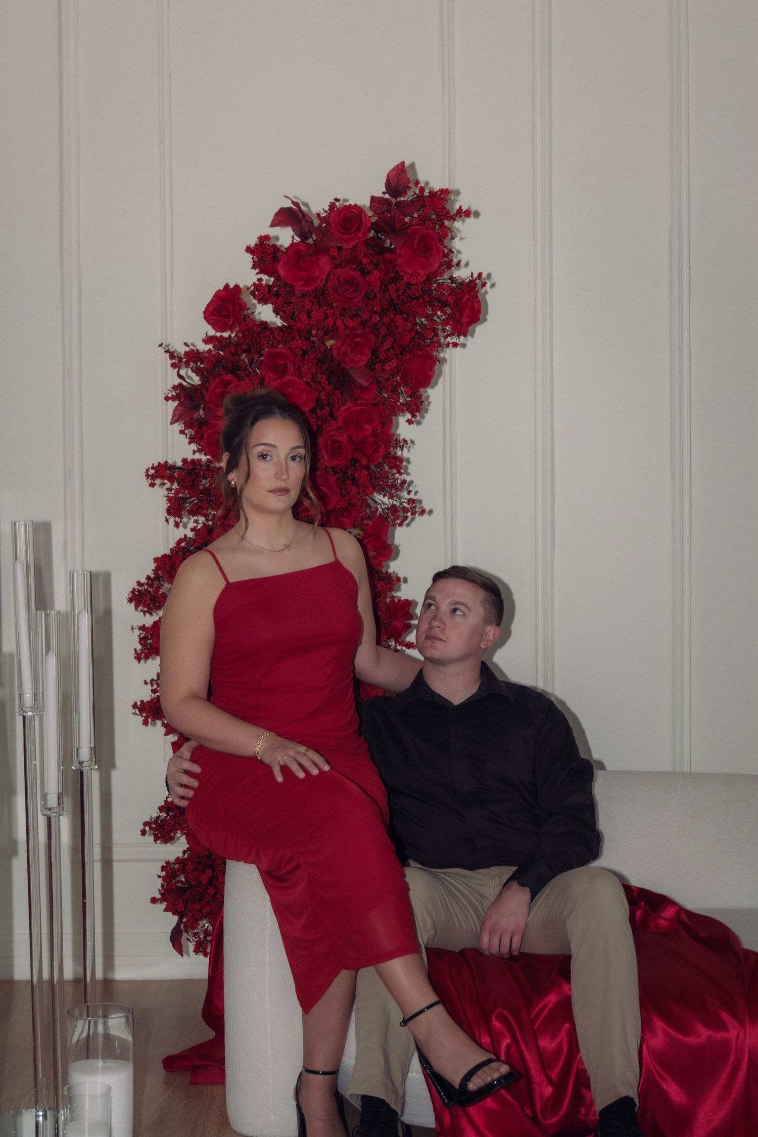 Couple poses elegantly with dramatic red rose floral arrangement in bright white minimalist interior.