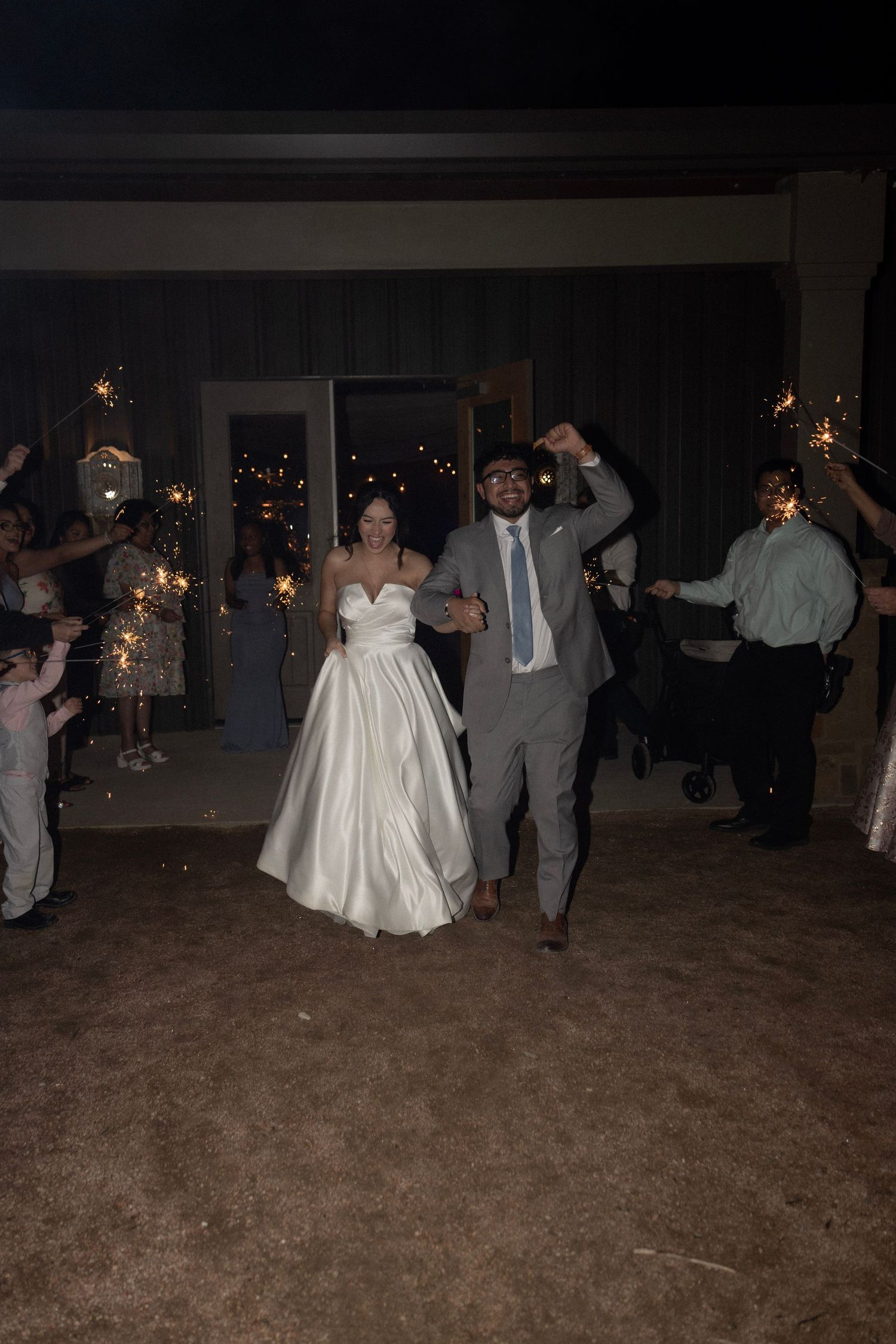 Newlyweds exit wedding reception through sparkler tunnel with guests celebrating at night outdoor venue.