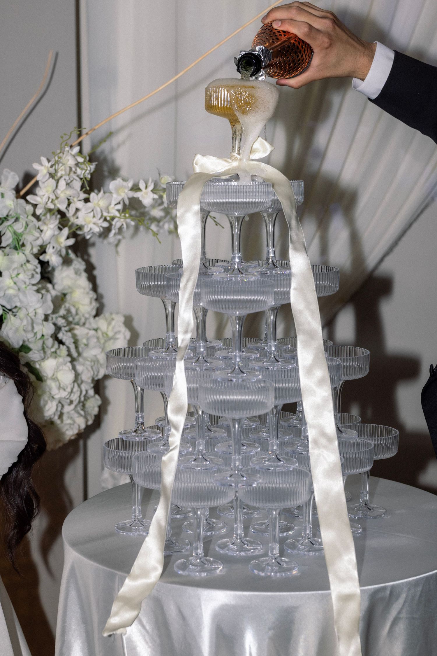 Champagne tower of coupe glasses with white ribbon being filled at elegant wedding reception.