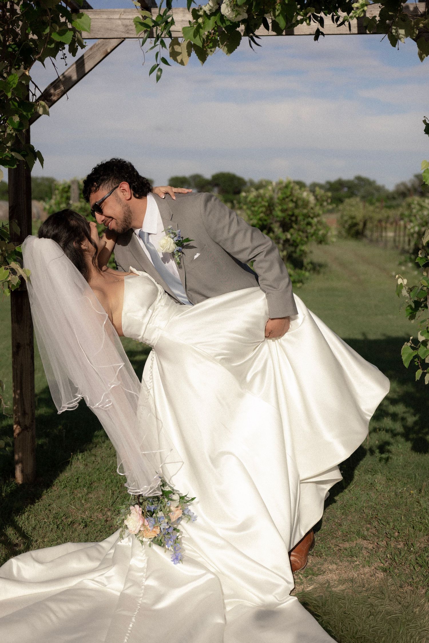 Groom lifts and kisses bride in white satin gown under wooden arch at vineyard wedding.