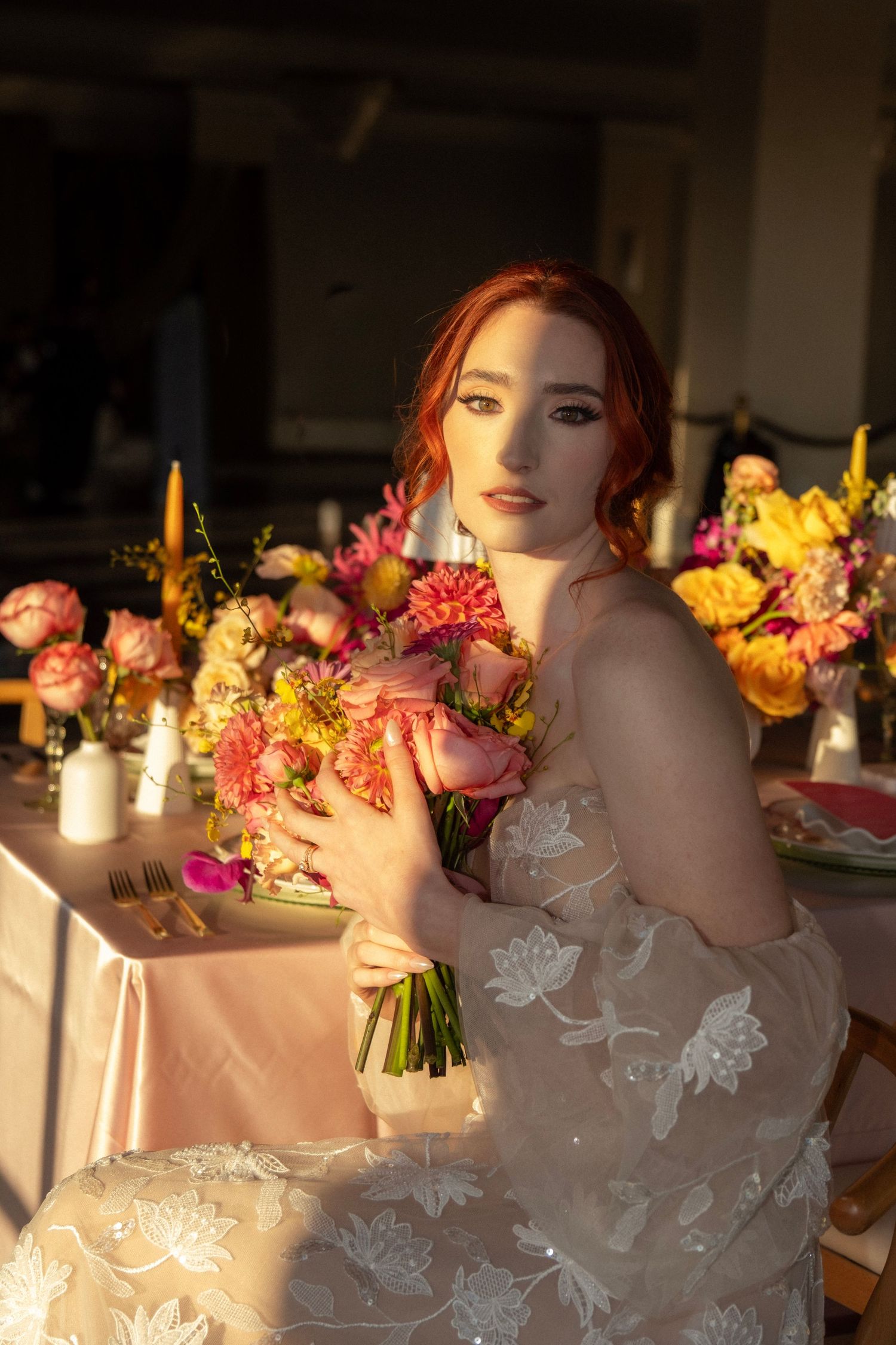 Beautiful red-haired bride in lace gown holding colorful bouquet at a floral wedding reception table.