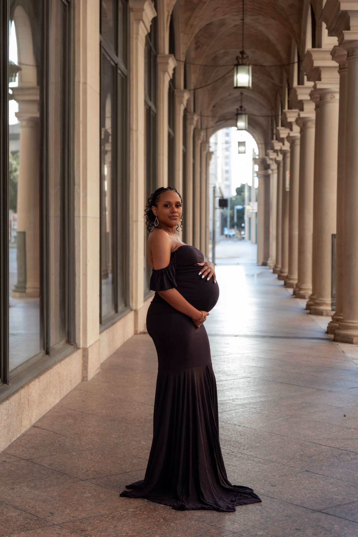 Pregnant woman in a dark off-shoulder gown posing gracefully in an arched colonnade hallway for maternity photos.