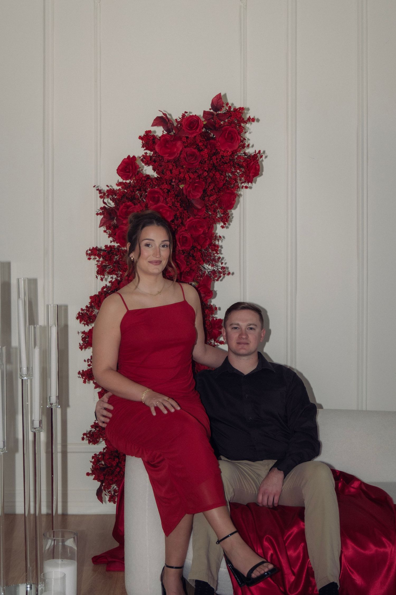 Couple seated together in white room with stunning red rose floral arrangement backdrop and candles.
