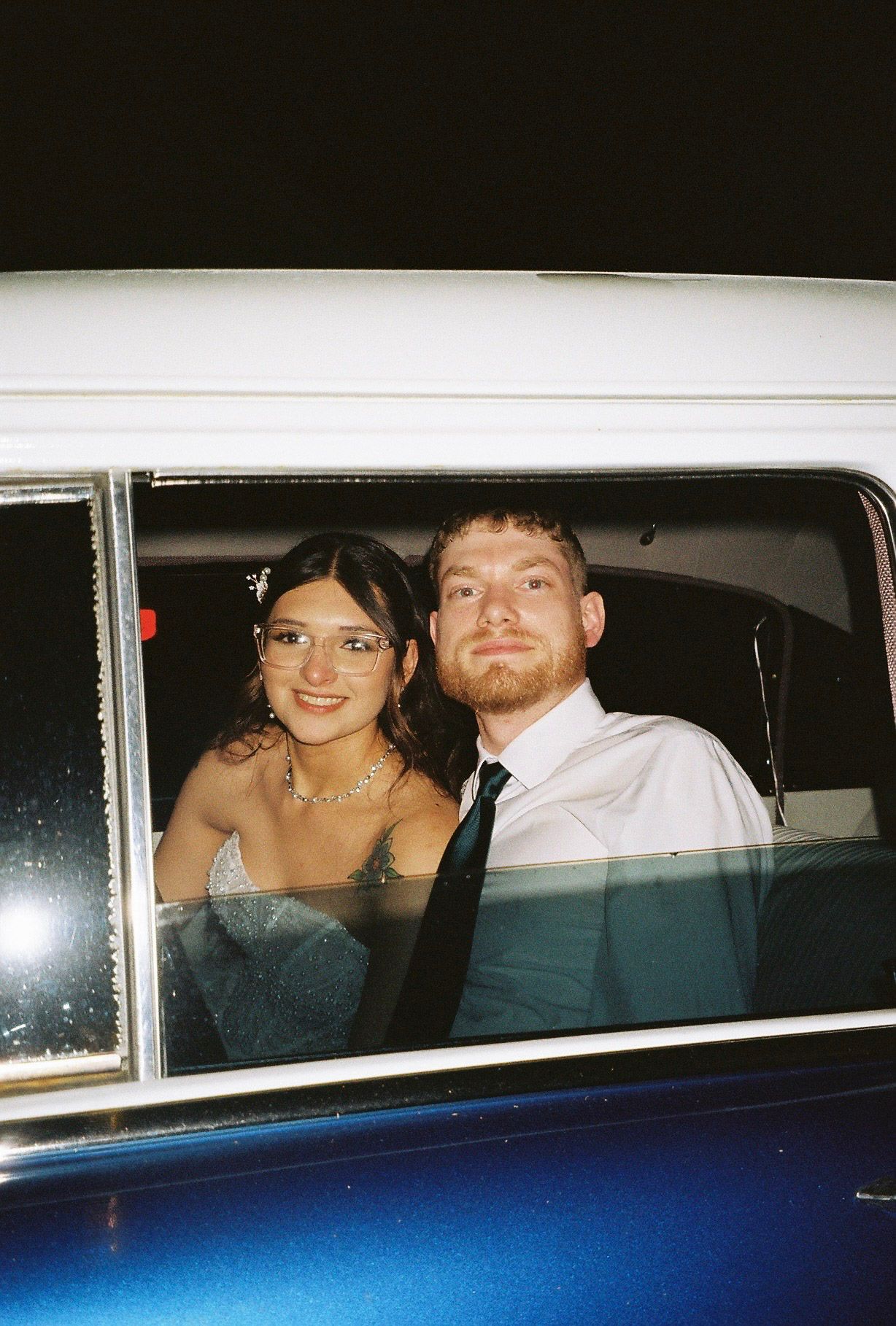 A smiling couple dressed formally poses together inside a vintage blue and white car at night.