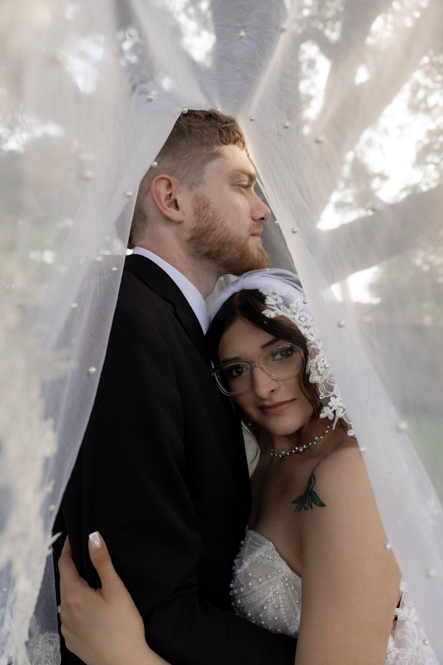 Bride and groom embrace under a sheer white veil, bride in lace headpiece and groom in black suit, outdoor wedding portrait.