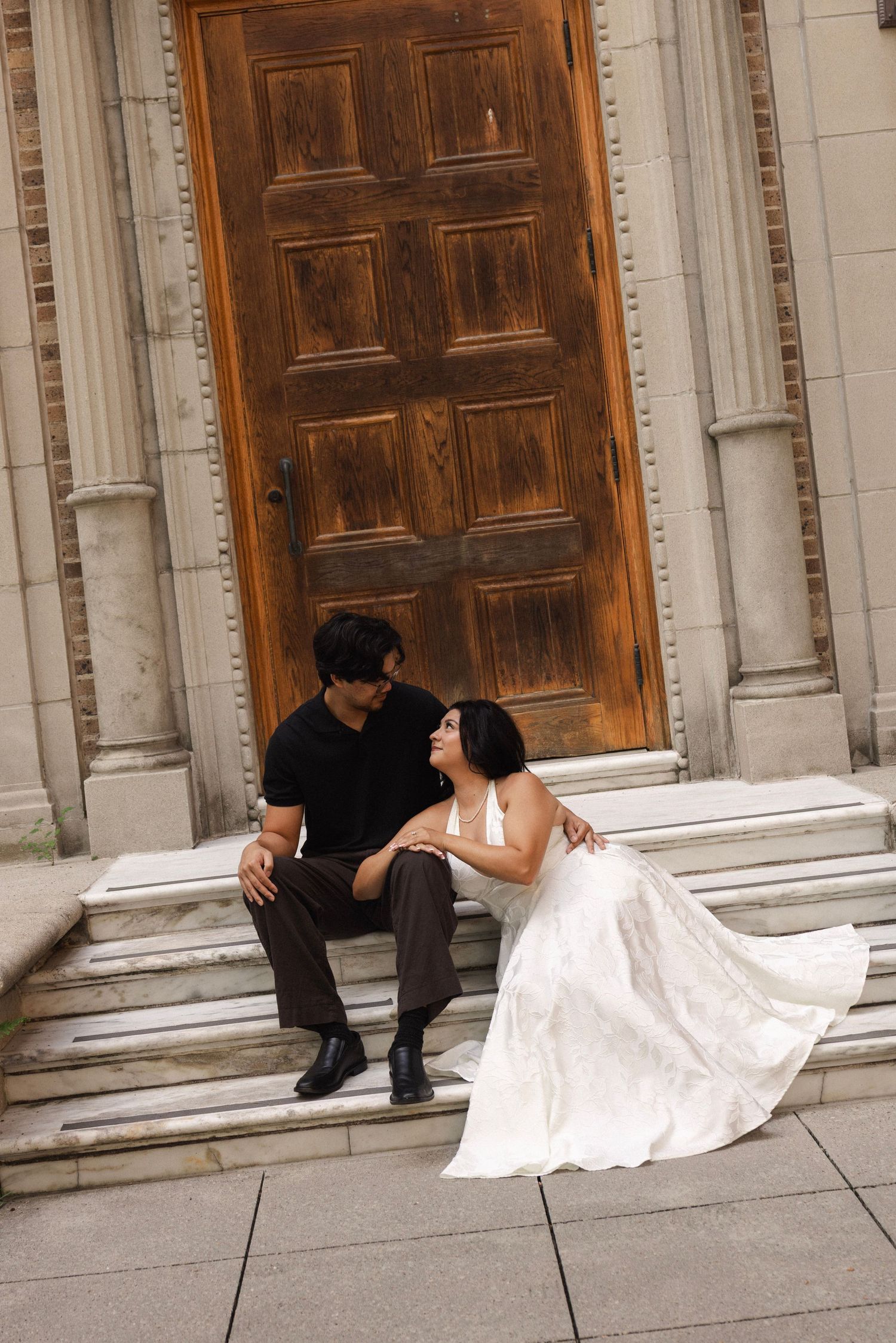 Couple in wedding attire sitting on stone steps before a large wooden door, sharing an intimate moment.