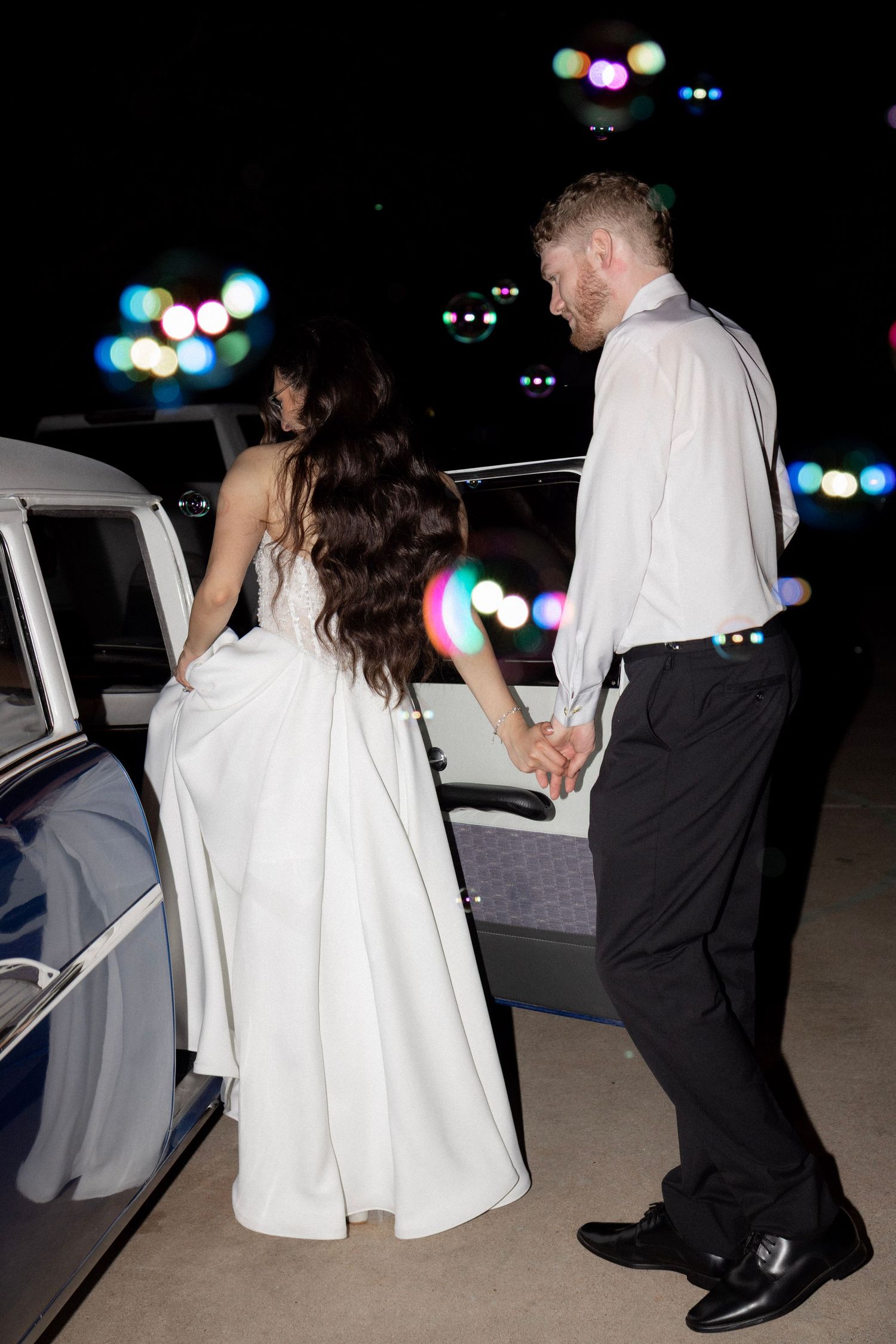 Bride and groom hold hands walking toward a vintage car at night.