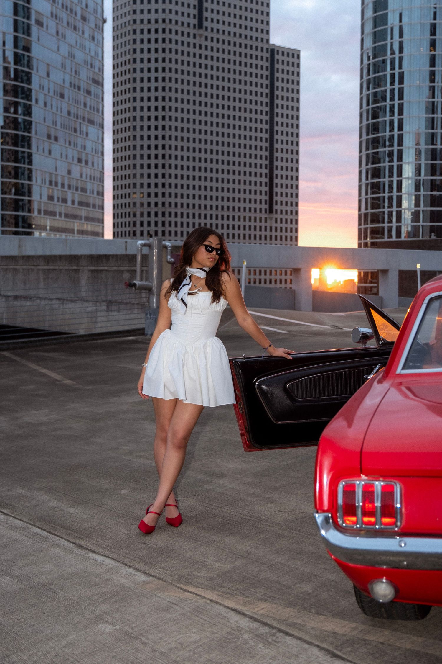 Woman in white dress posing next to red classic Mustang on rooftop parking garage at sunset with city skyline.