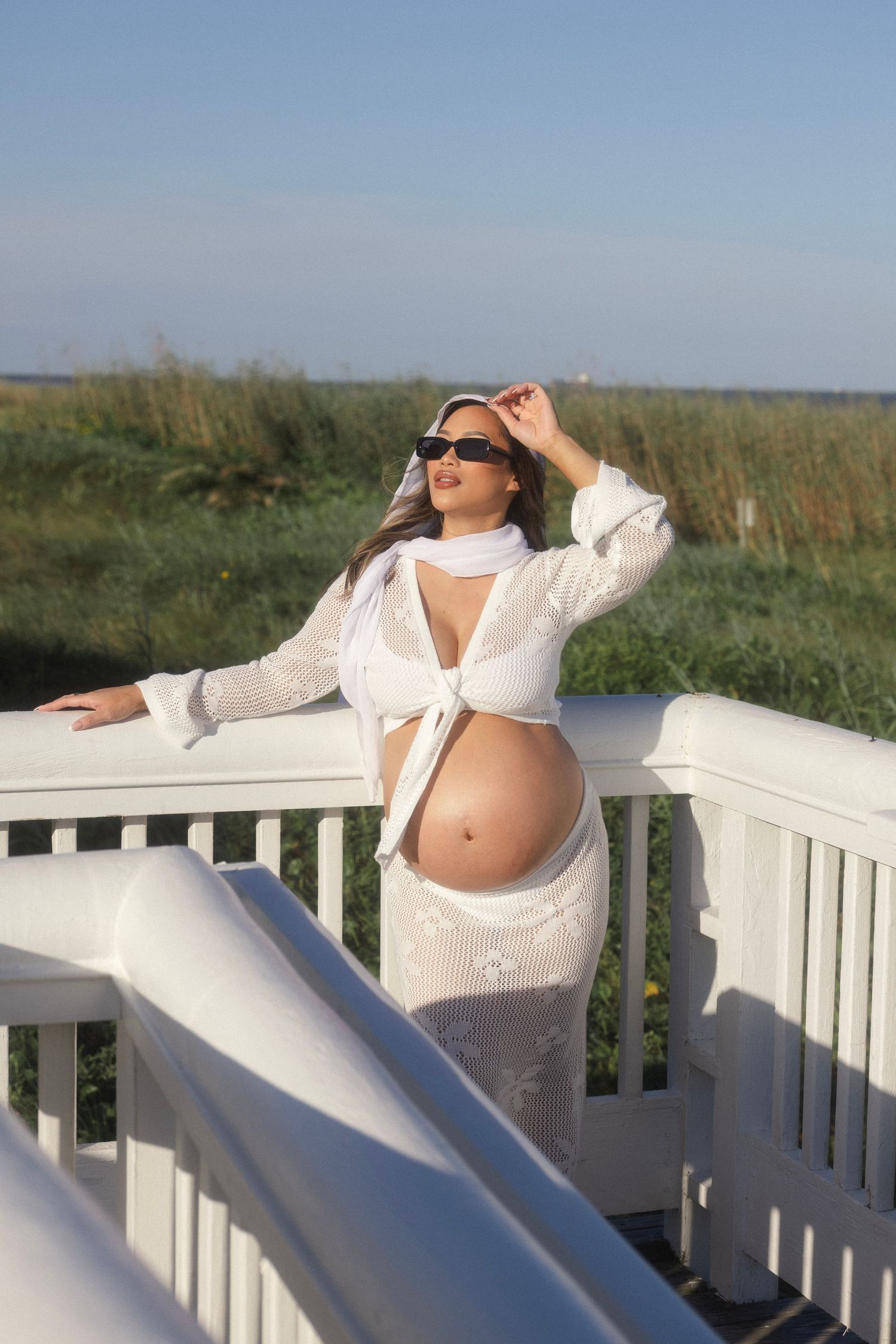 Pregnant woman in white tied crop top and skirt poses on white railing outdoors wearing sunglasses.