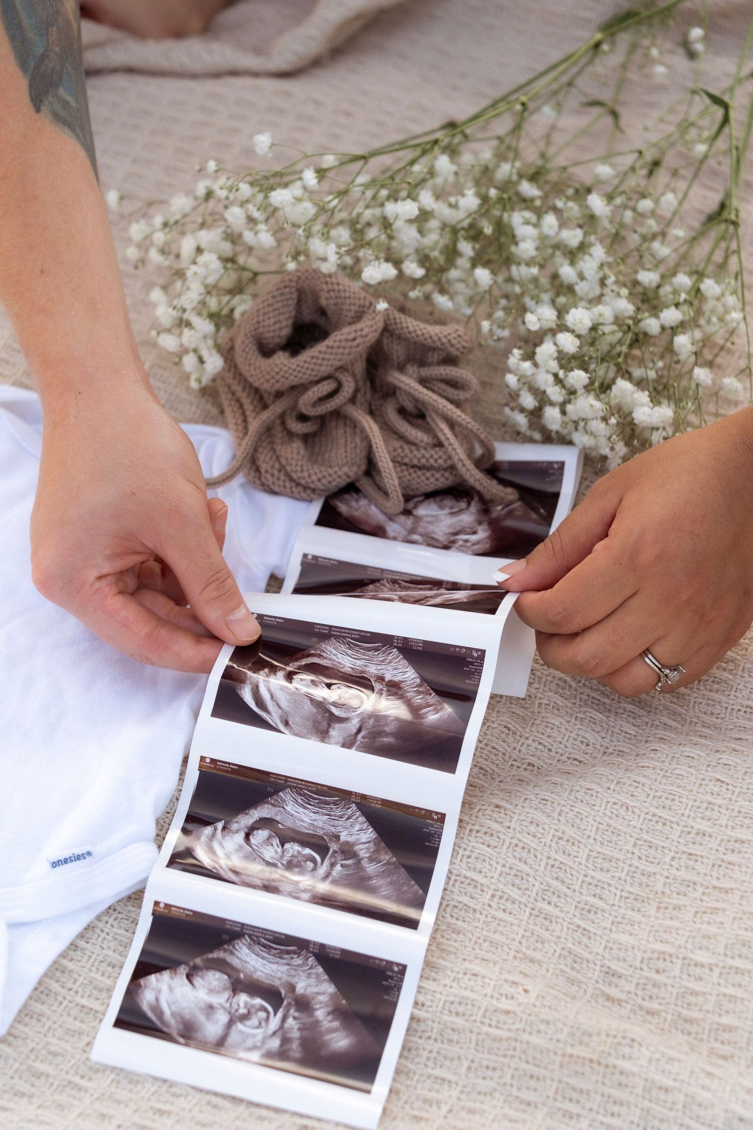 Couple holding ultrasound photos with baby booties and white flowers on a neutral linen background.