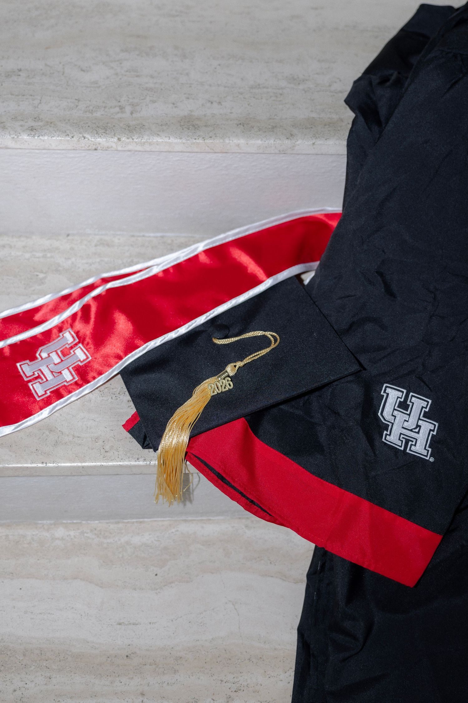 University of Houston black graduation cap, gown, and red satin honor stole on steps.