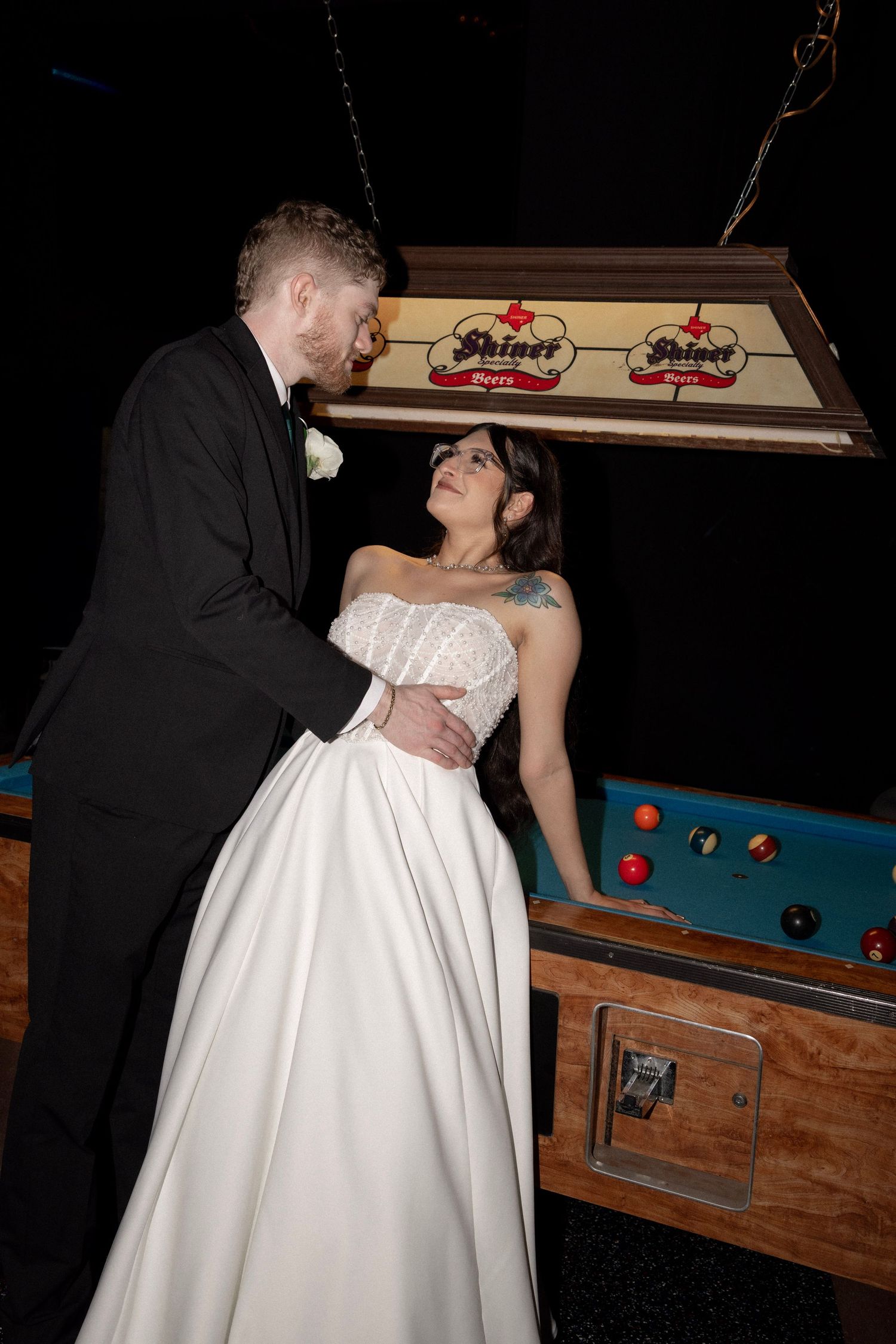 Bride and groom share a romantic moment beside a Shiner Beer pool table light at wedding reception.