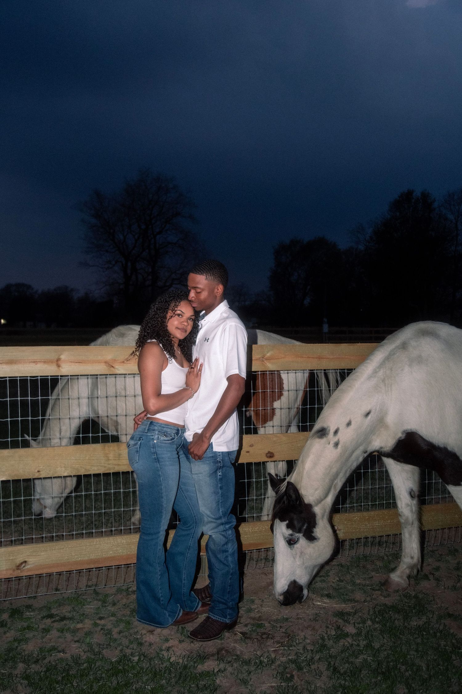 Couple embracing at night by a wooden fence with white horses grazing nearby on a farm.