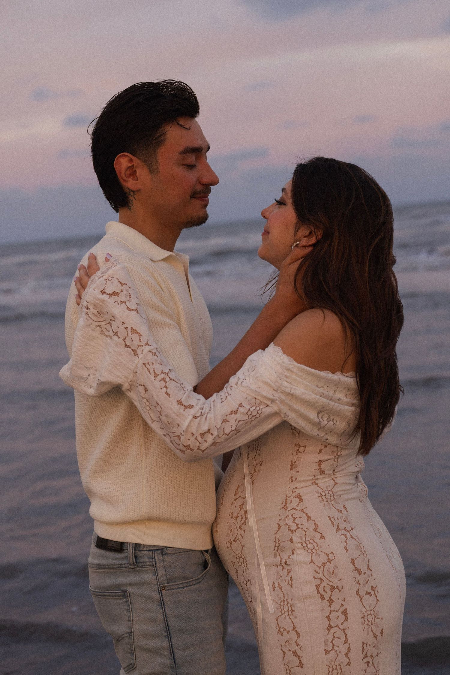 Couple embracing romantically on a beach at sunset, woman in white lace off-shoulder dress, man in cream polo and jeans.