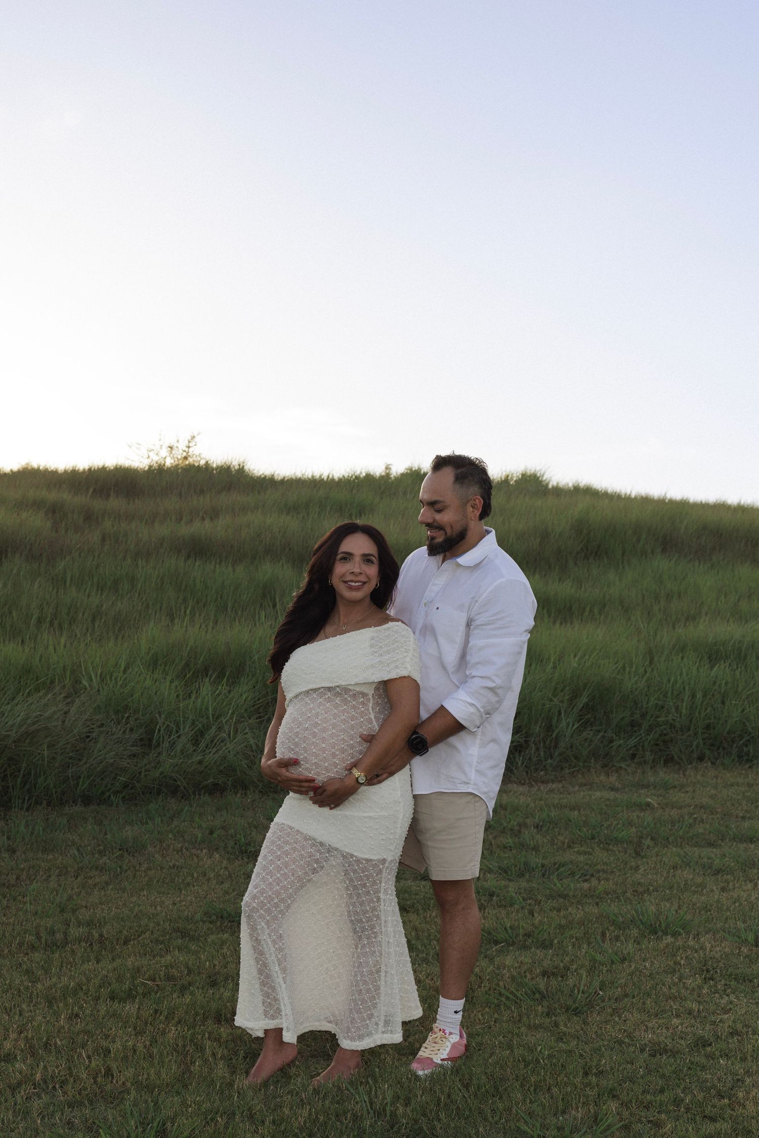 Couple posing outdoors in white outfits during maternity photo shoot on green grassy hill at dusk.