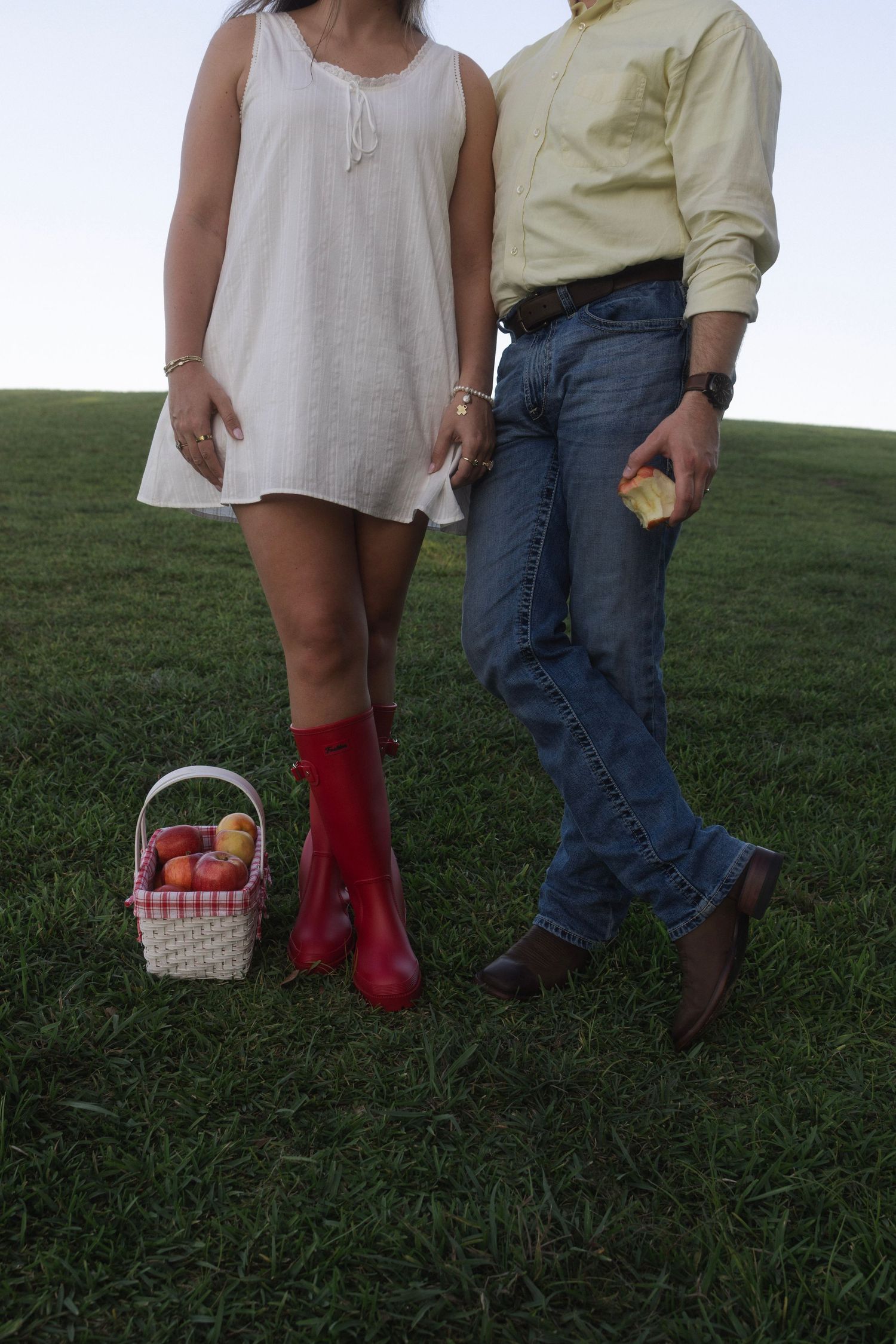 Couple standing on green grass with a wicker basket of apples, woman in red boots and white dress, man in jeans.