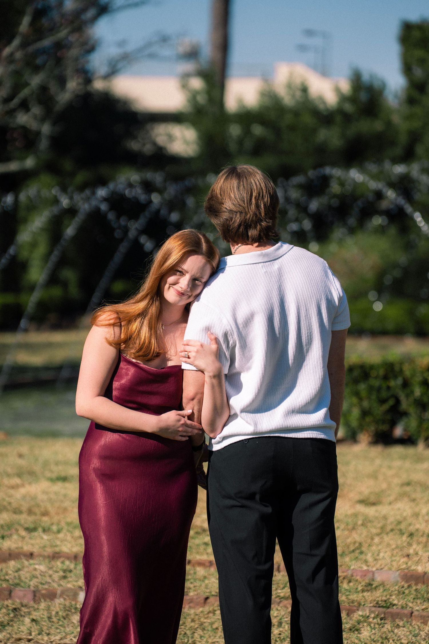 Young couple in formal wear holding hands by a garden fountain, woman in burgundy satin dress smiling warmly.