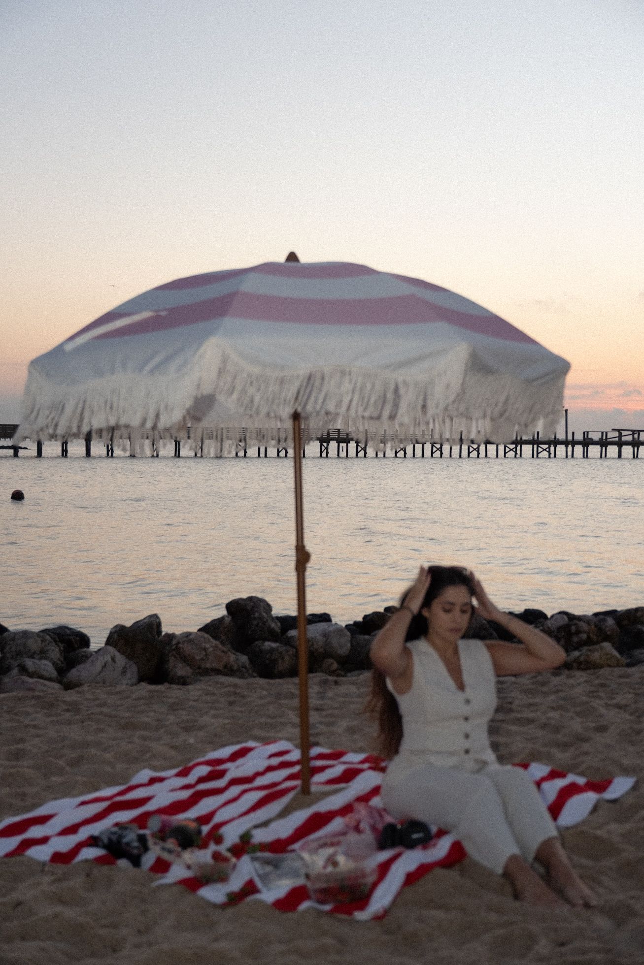 Woman sitting on striped beach blanket under fringed umbrella at sunset by the ocean pier.