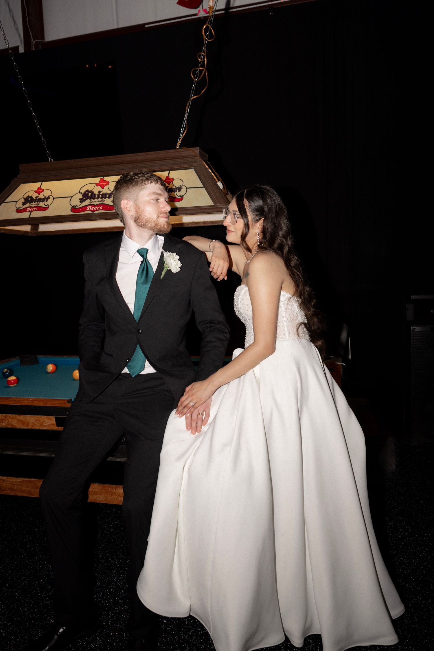 Bride and groom pose together near a pool table under a Shiner beer lamp.