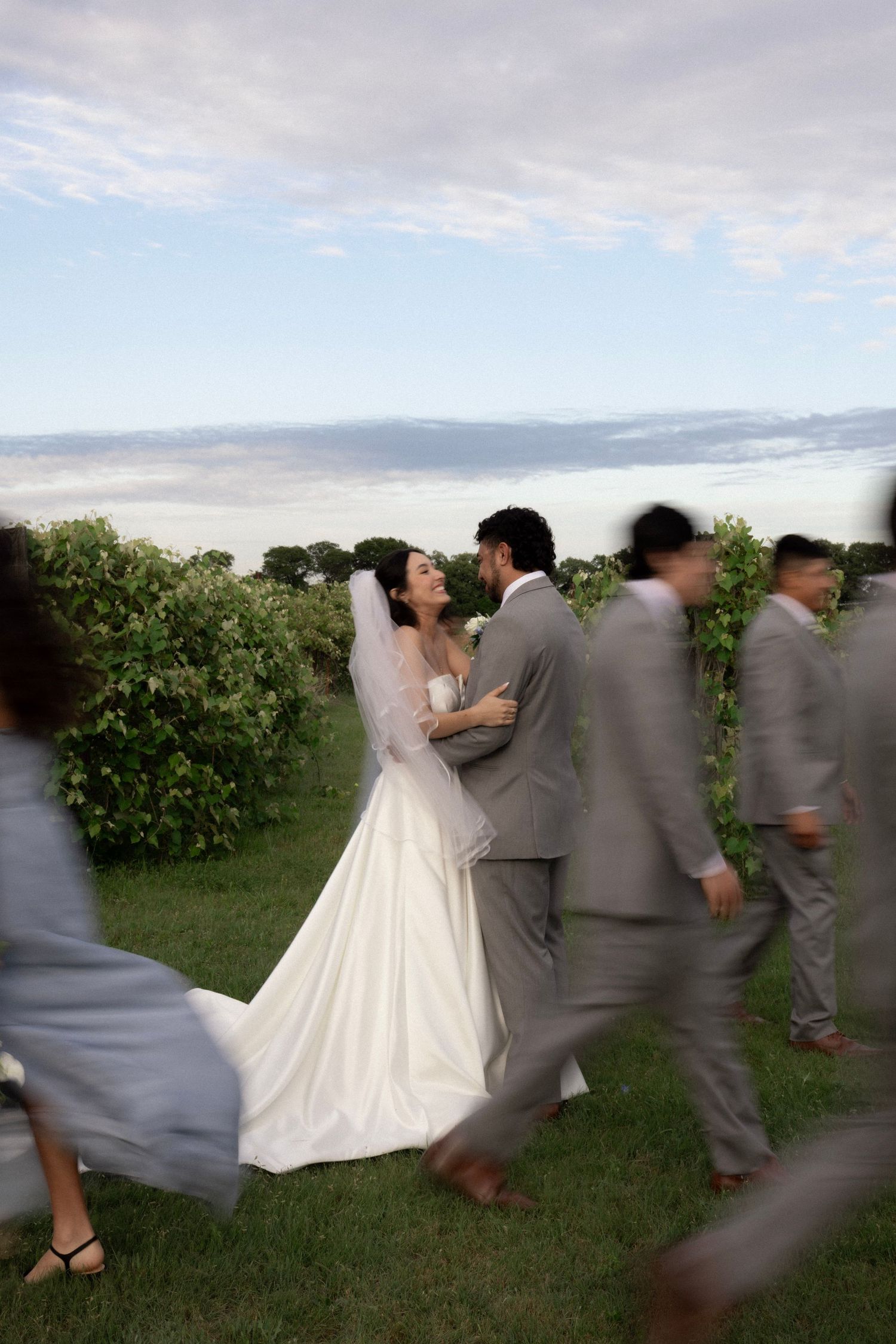 Bride and groom share intimate moment surrounded by blurred wedding party walking past vineyard backdrop.