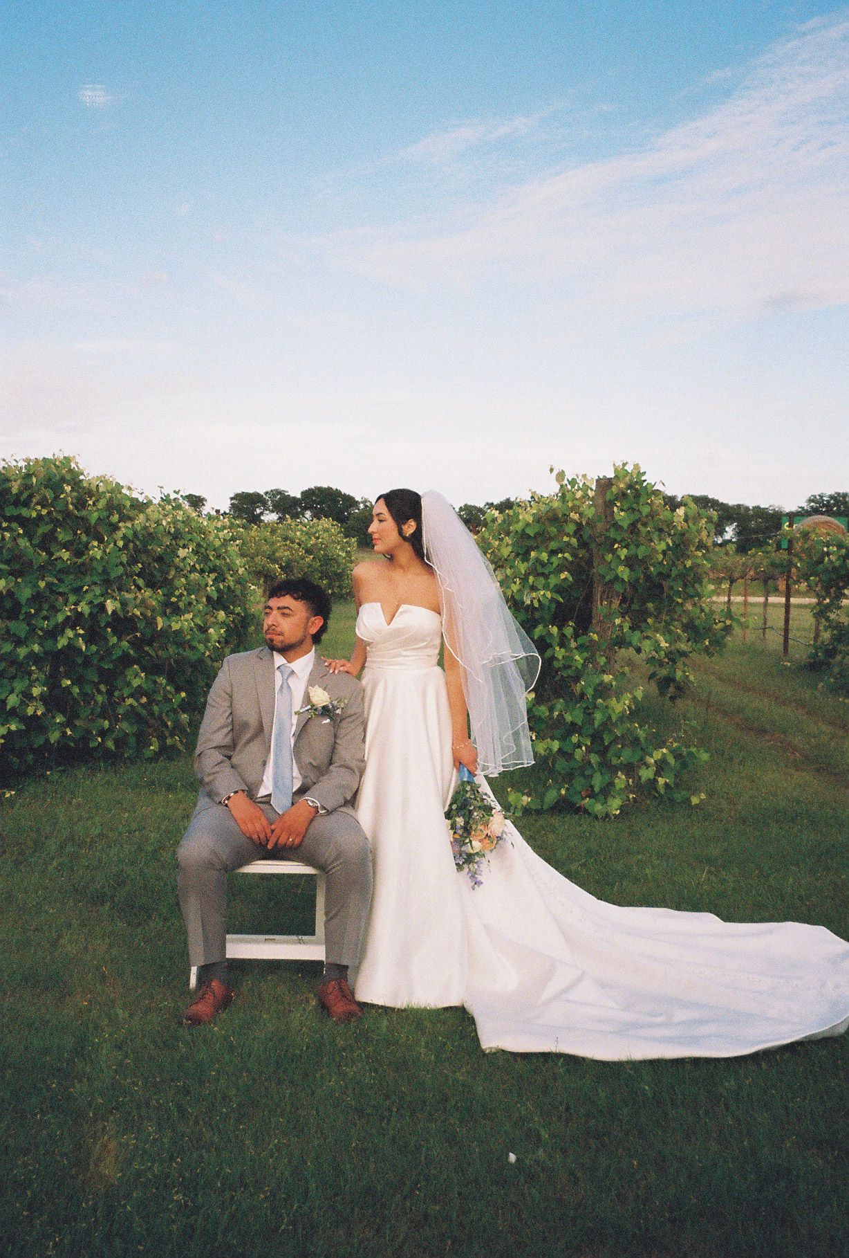 Bride and groom pose in a lush vineyard, bride in white gown with veil, groom in gray suit.